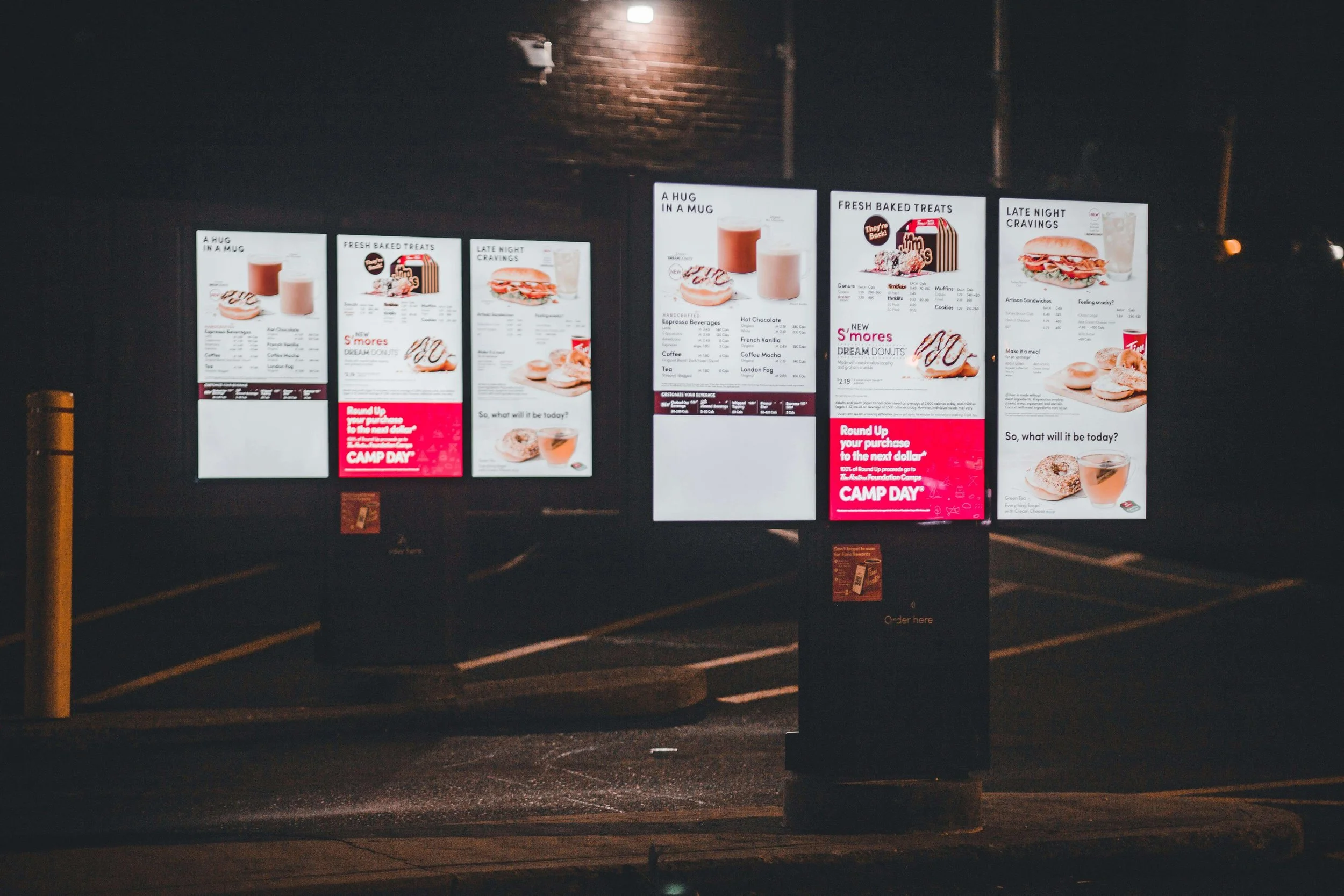 Outdoor LED digital menu panels illuminating a fast-food drive-thru at night, displaying vibrant food promotions and menu items