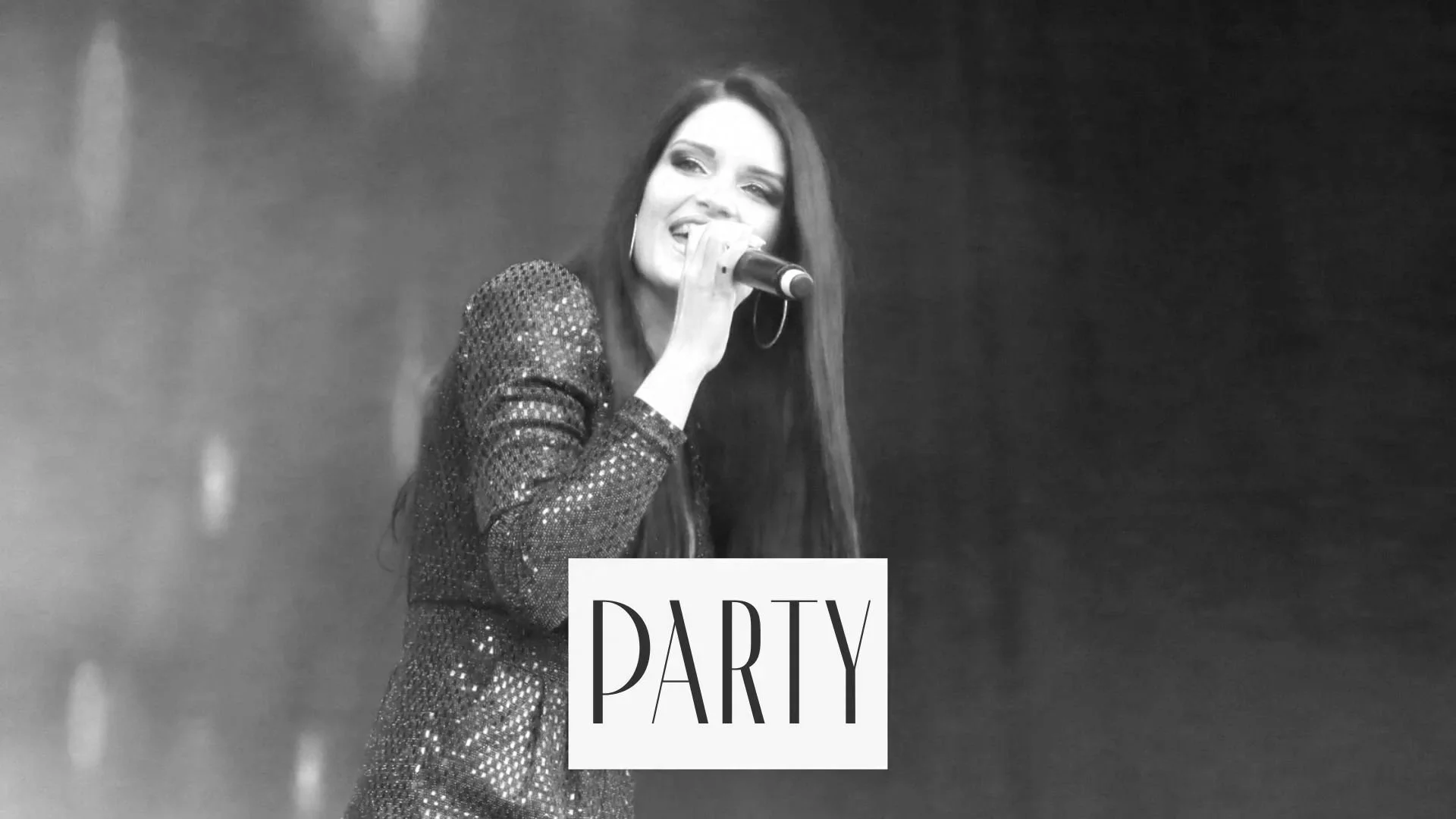 A woman with long dark hair, wearing a sparkly jacket, holding a microphone, smiling and singing on stage at a party.