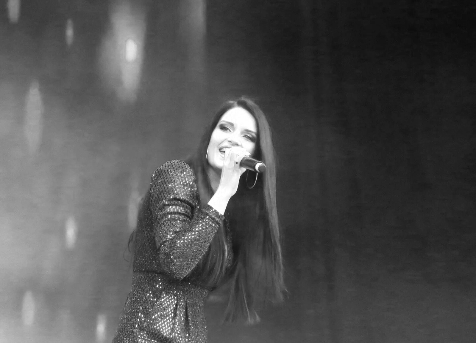 Black and white photo of a woman singing into a microphone, wearing a sparkly long-sleeve dress, with long hair and hoop earrings, smiling while performing on stage.