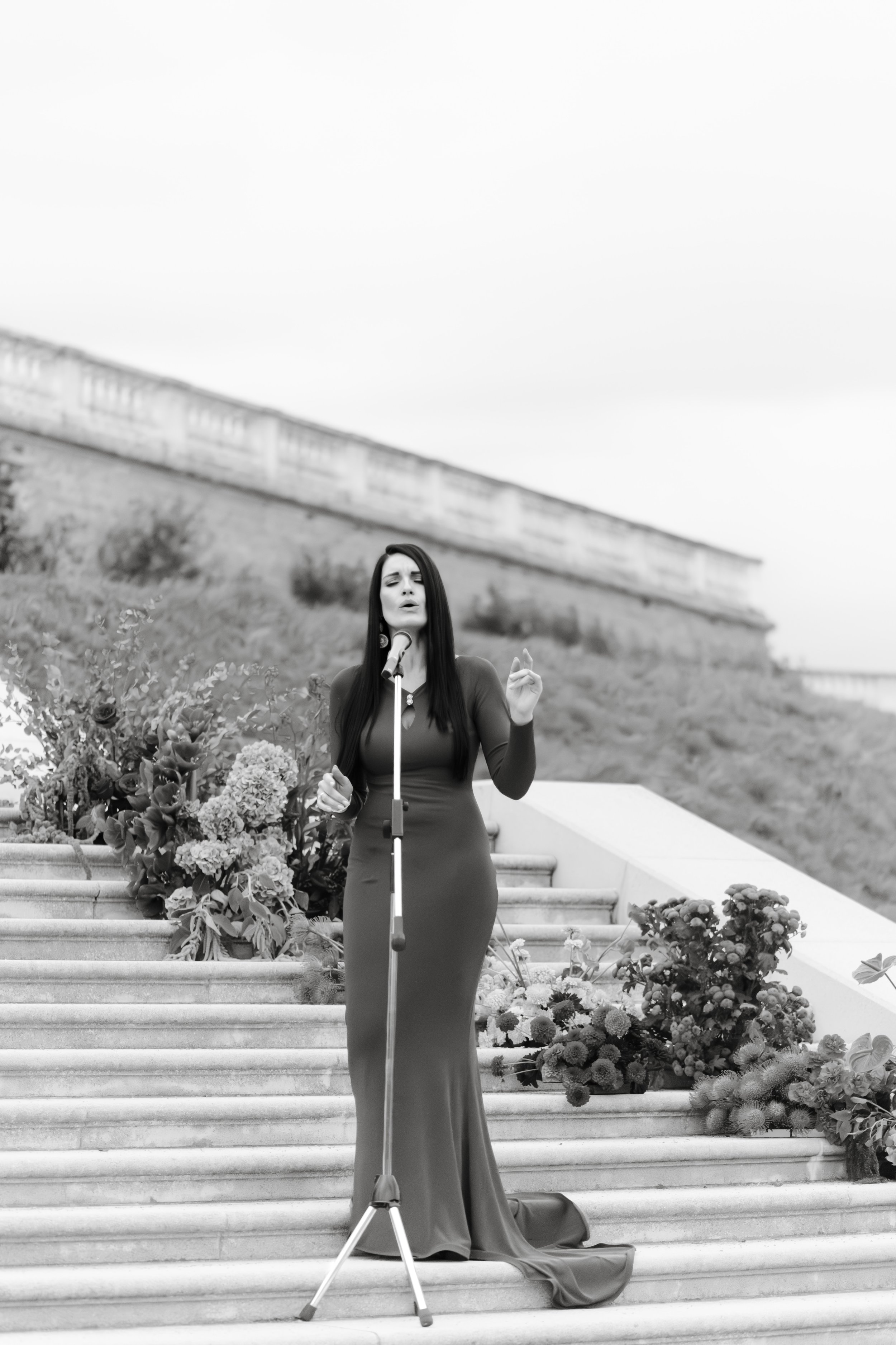 A woman singing on outdoor stairs decorated with flowers, wearing a long dress.