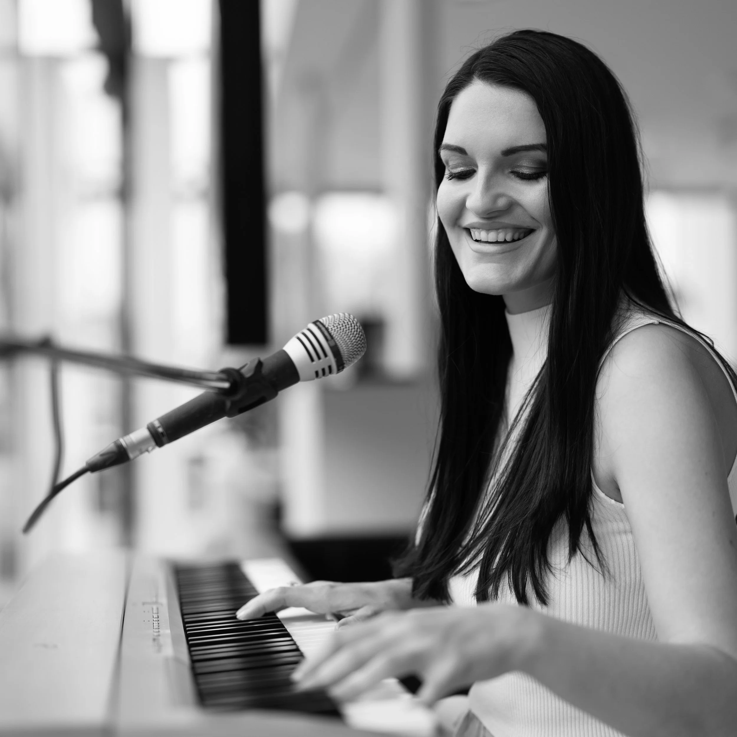 A woman with long dark hair playing piano and singing into a microphone, smiling with eyes closed in a cozy indoor setting.