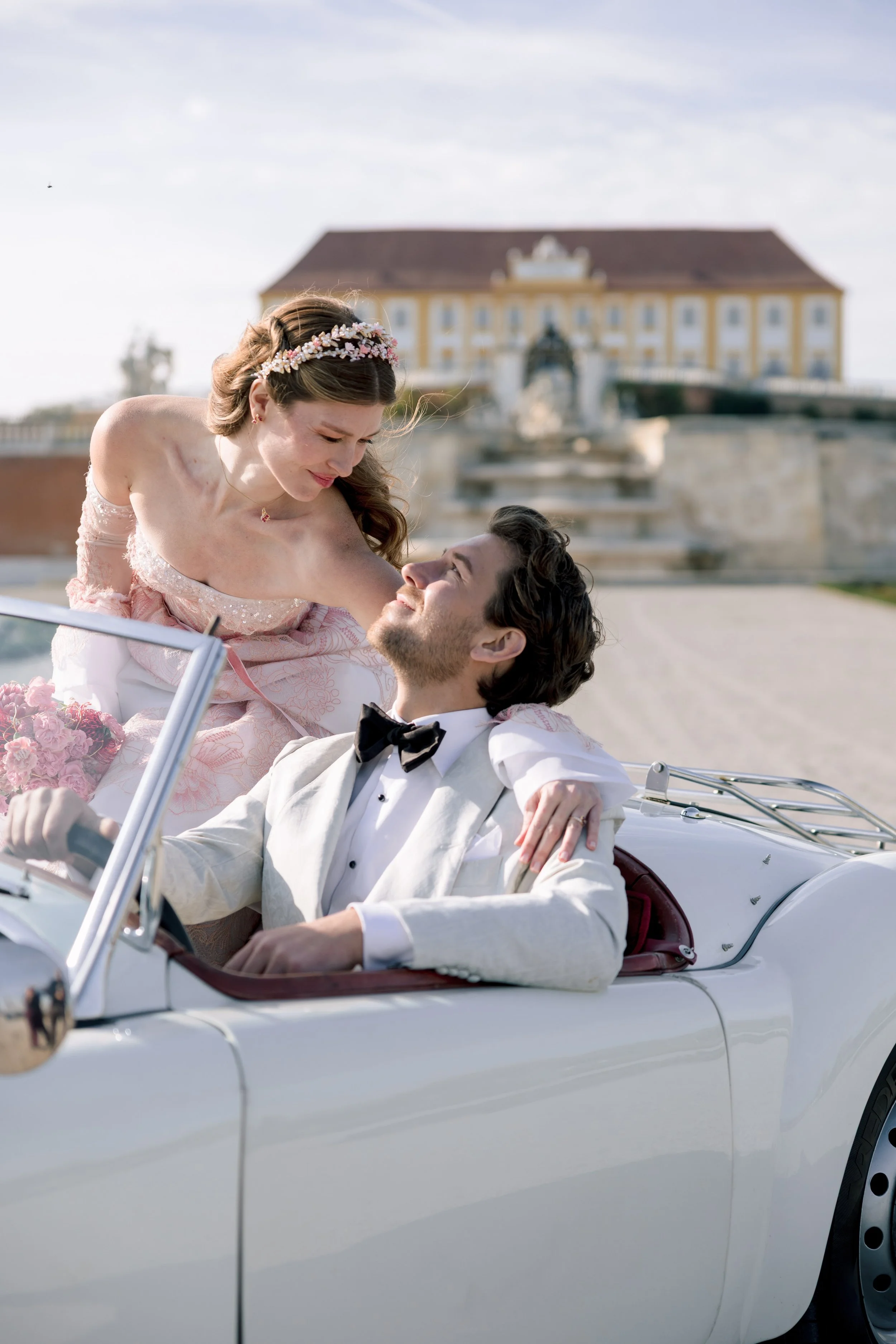 A couple in wedding attire sharing a joyful moment in a vintage white convertible car, with a grand castle in the background. Luxury destination weddings in Austria. Photo taken by Sotiris Tsakanikas.