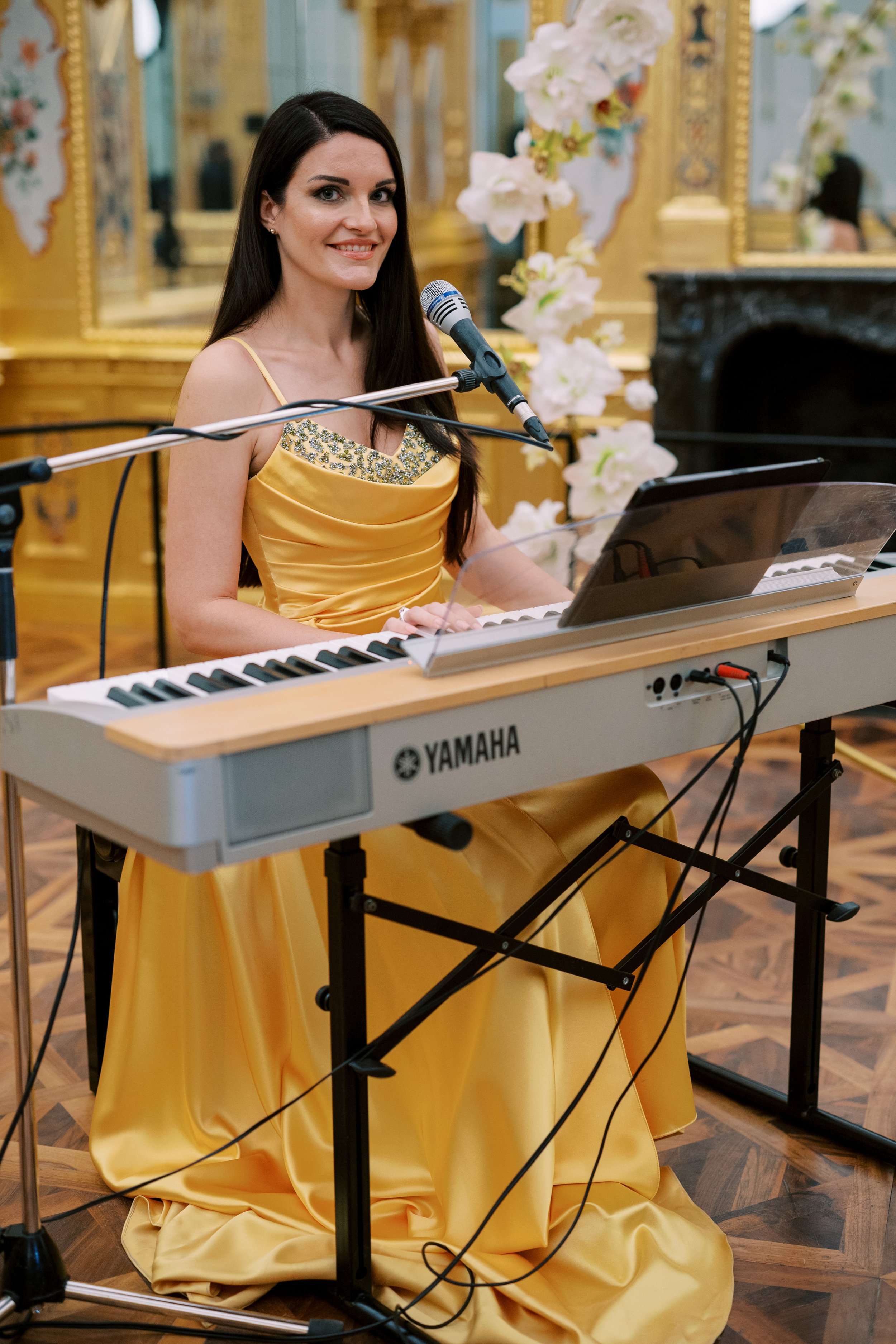 A woman in a yellow satin gown sitting at a Yamaha keyboard, smiling, with a microphone, in a decorated room with flowers and gilded mirrors.