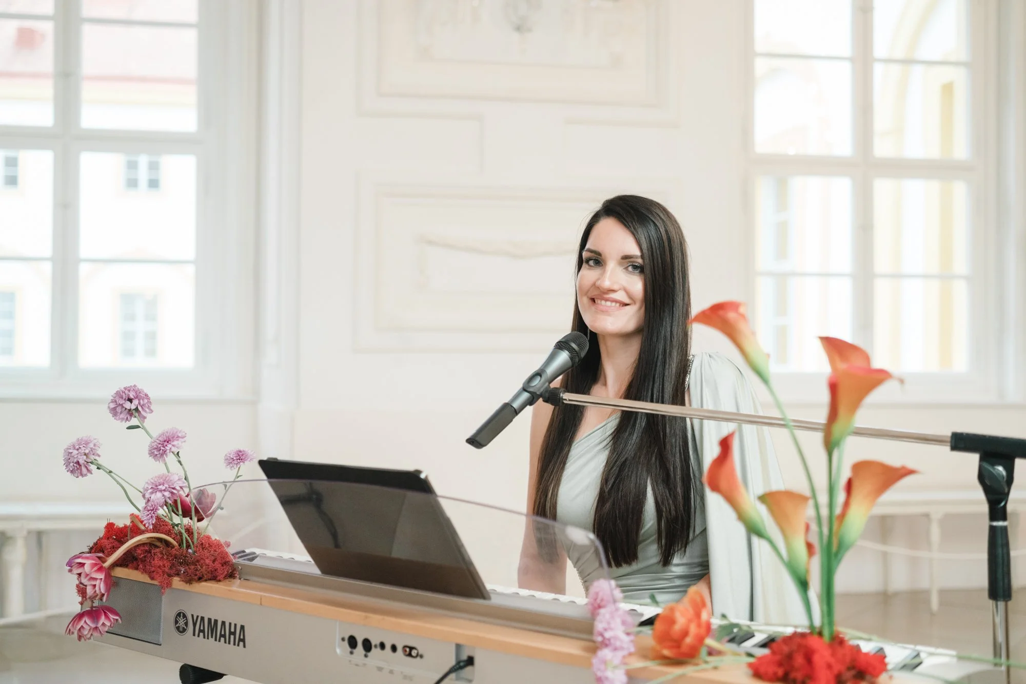 A woman with long dark hair sitting at a Yamaha keyboard with a microphone, surrounded by pink and orange flowers in a bright room with large windows.