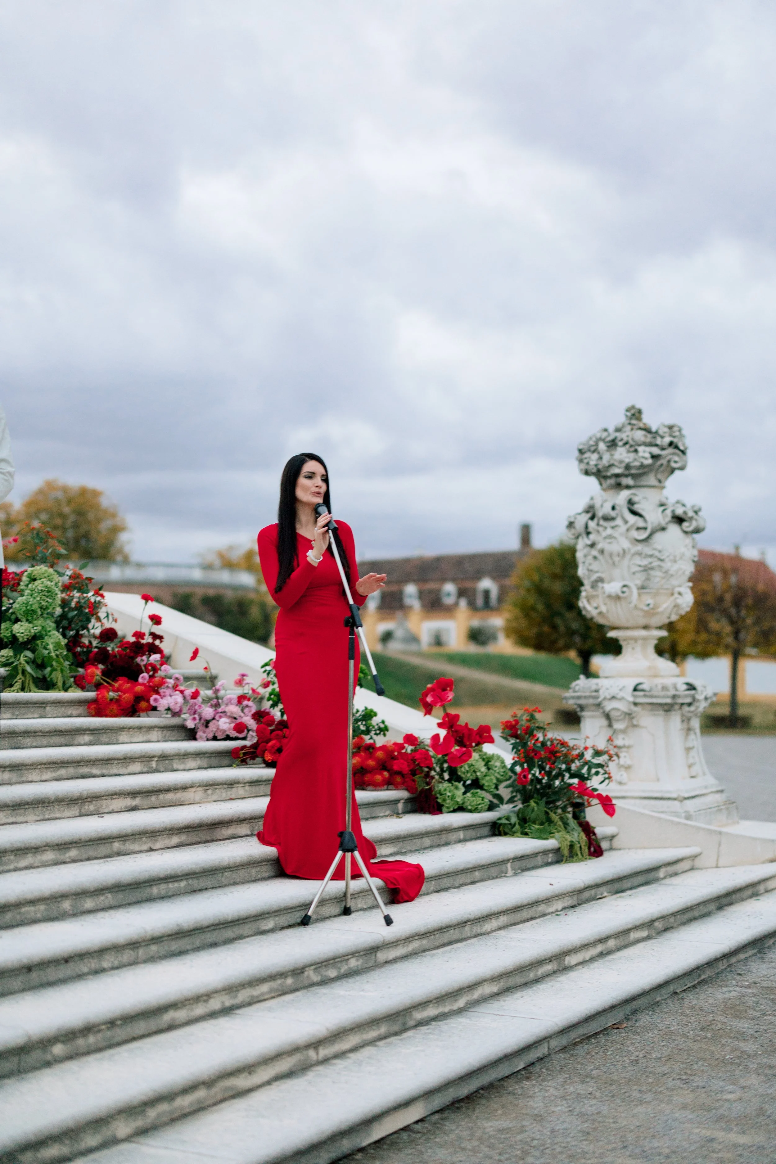 Woman in a long red dress speaking into a microphone on outdoor steps decorated with flowers, with cloudy sky and historic buildings in the background.