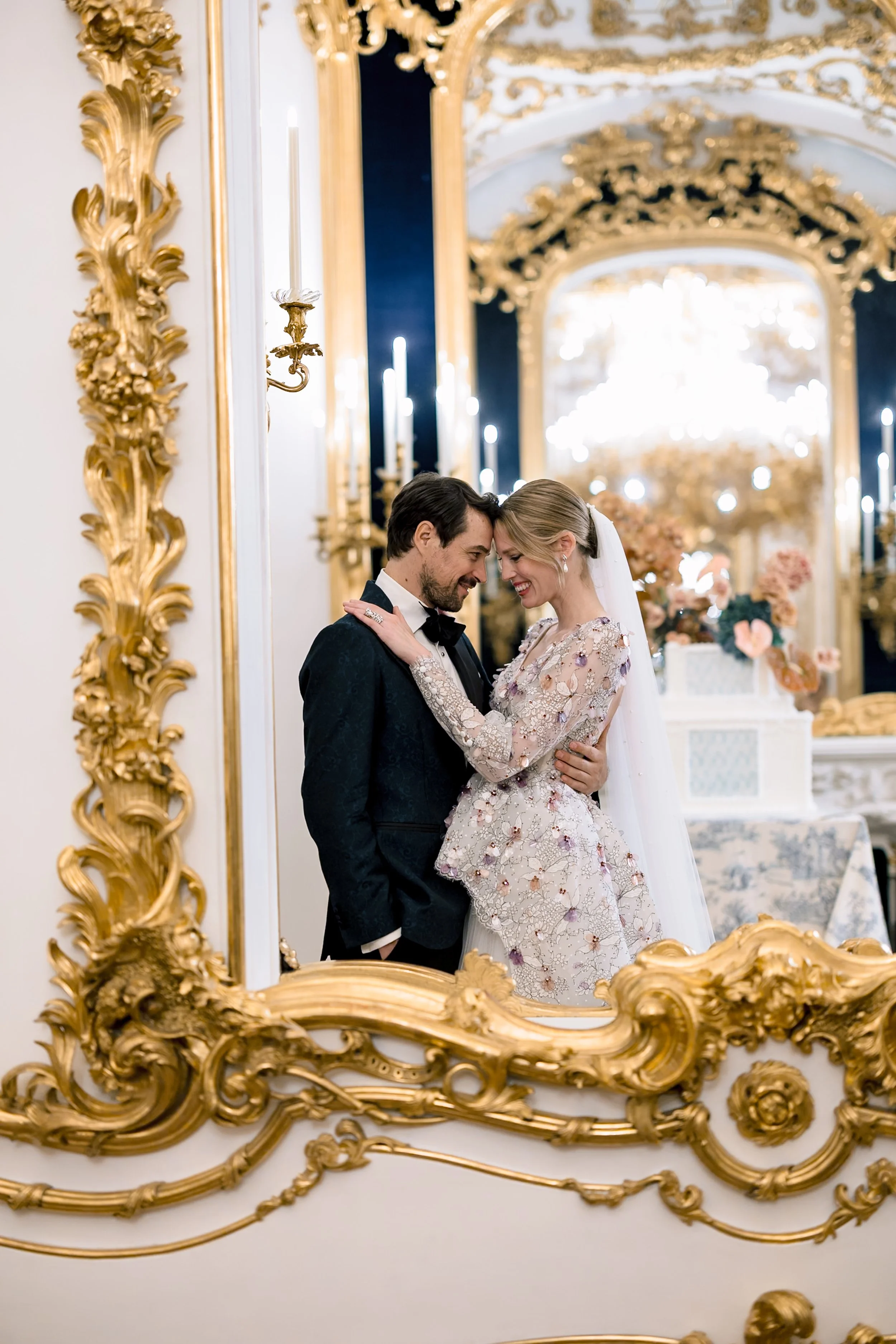 A bride and groom smiling and touching foreheads in front of a large, ornate gold-framed mirror at an elegant, luxurious wedding reception with gold and floral decor and a wedding cake in the background. Photo by Sotiris Tsakanikas.