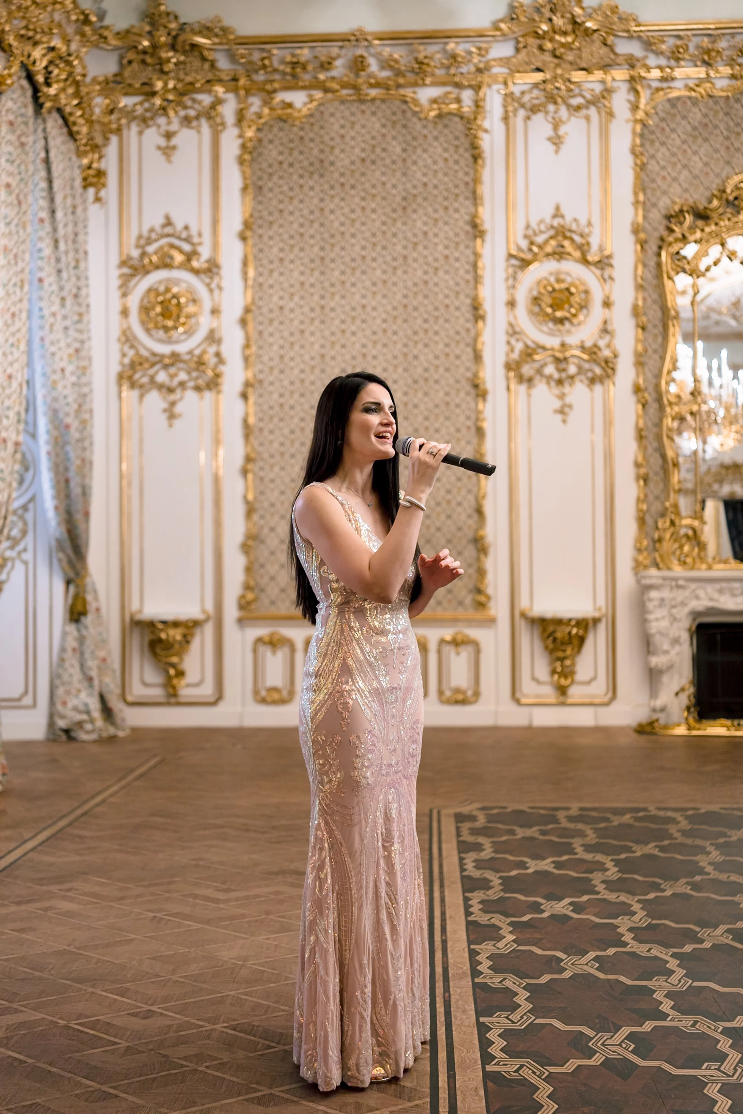 A woman in an elegant, gold and pink gown singing into a microphone in a luxurious, ornate room.