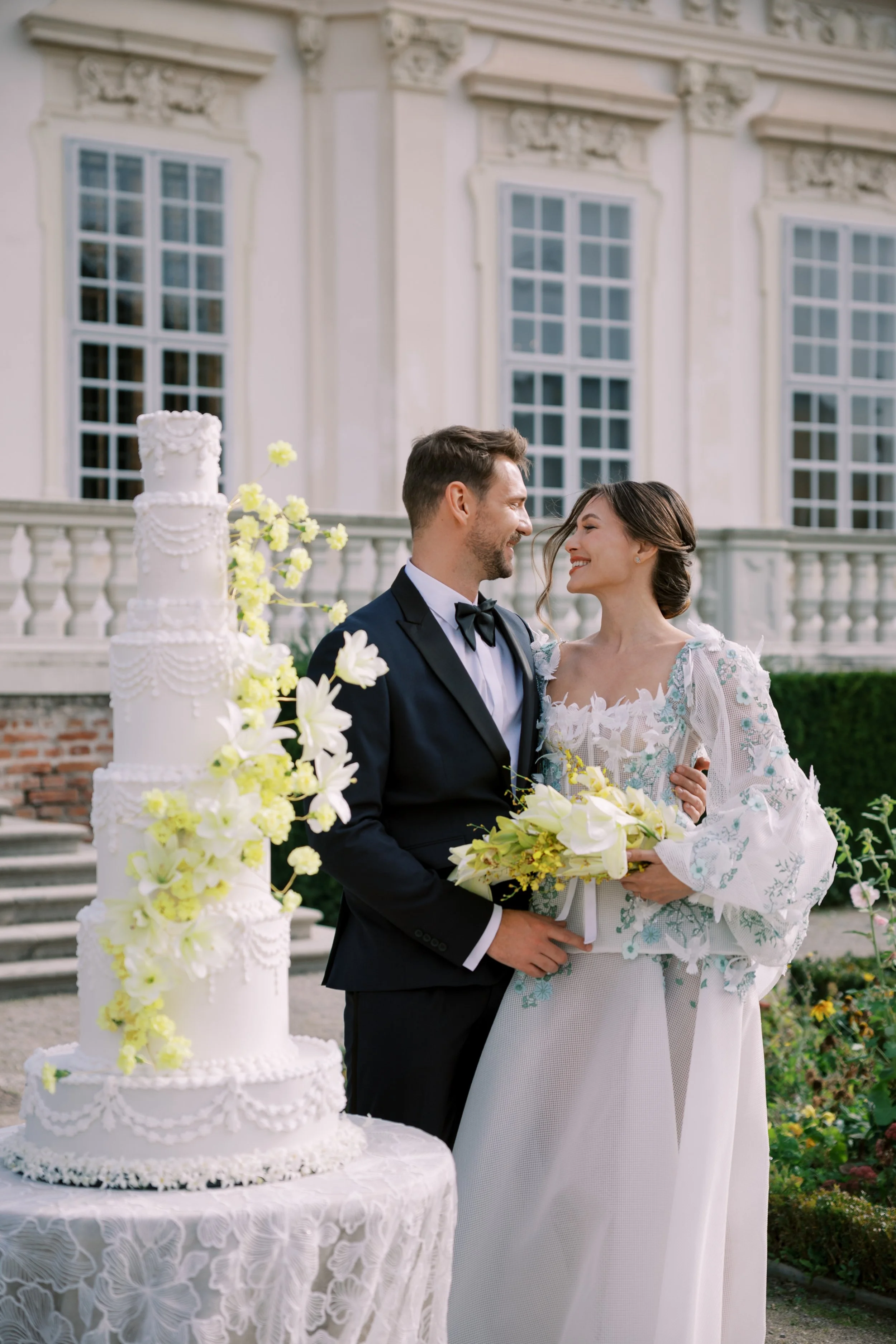 A bride and groom smiling at each other next to a tall white wedding cake decorated with flowers, outdoors in front of a historic building with large windows.