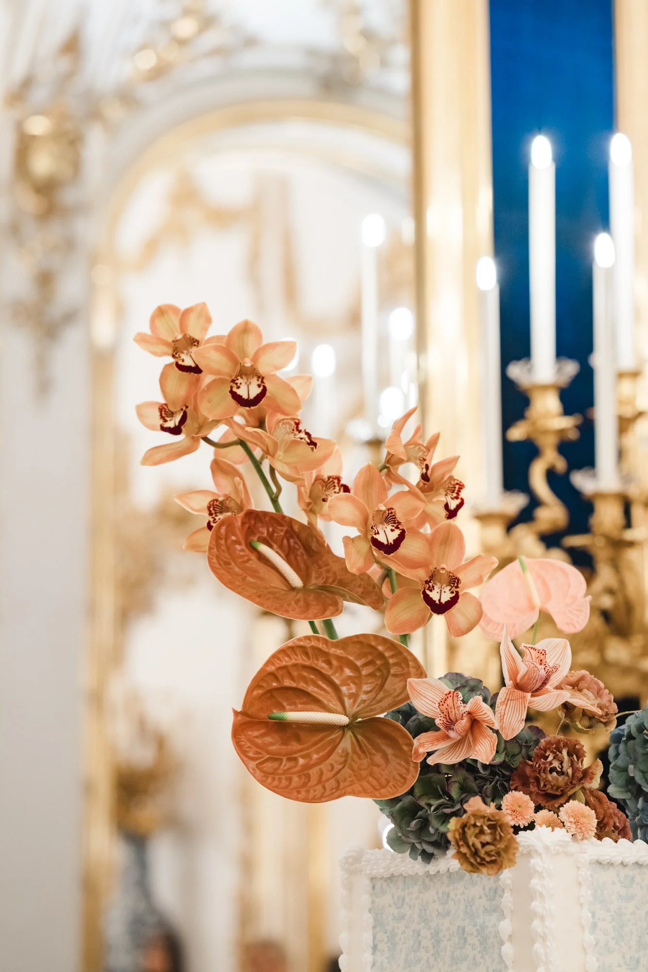 Flower arrangement with pink orchids, orange anthuriums, and other flowers in a decorative vase on a table in a ornate room with chandeliers and candle holders.