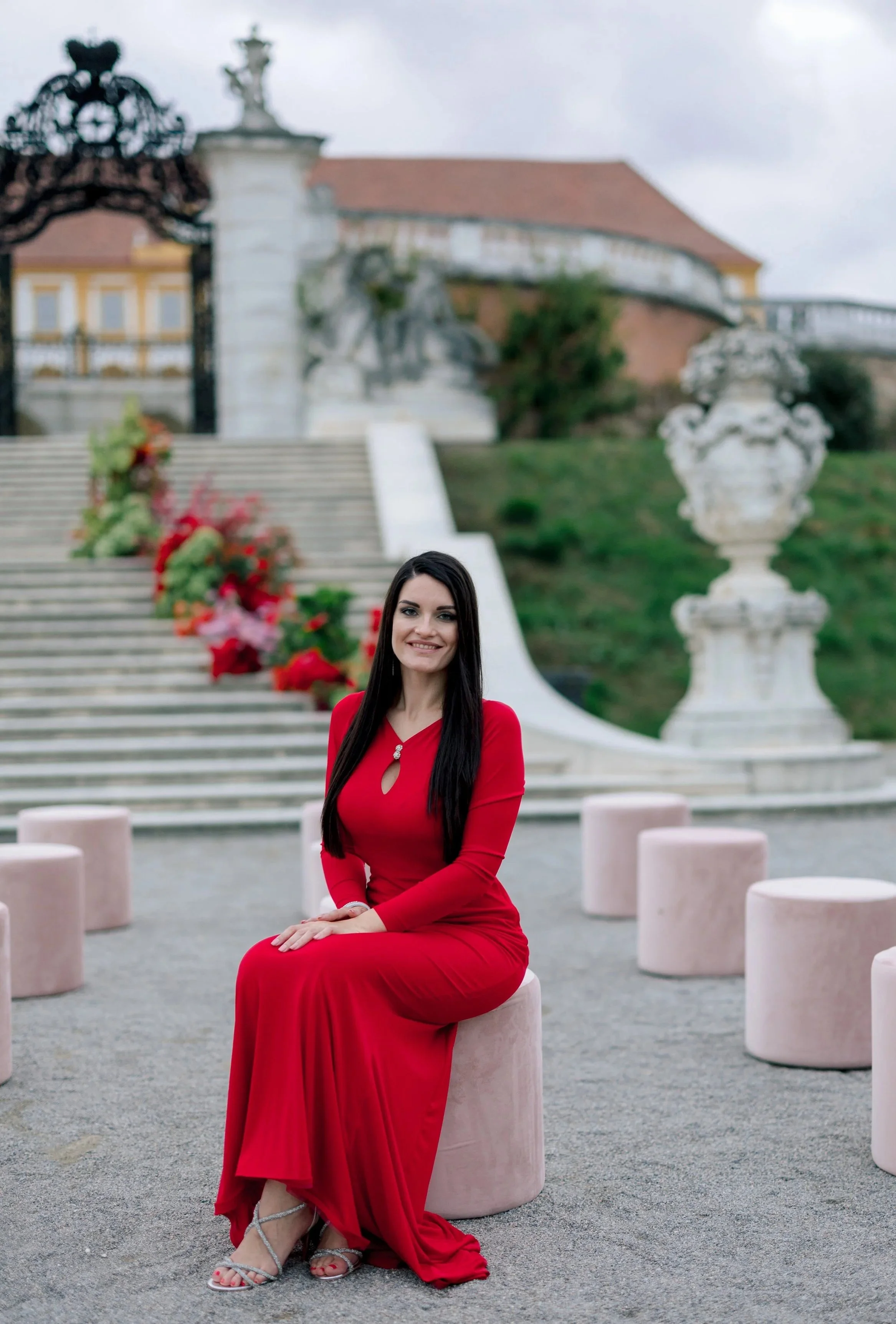A woman in a long red dress sitting on a pink ottoman outdoors, with a decorative stairway, sculptures, large urns, and a historic building in the background.
