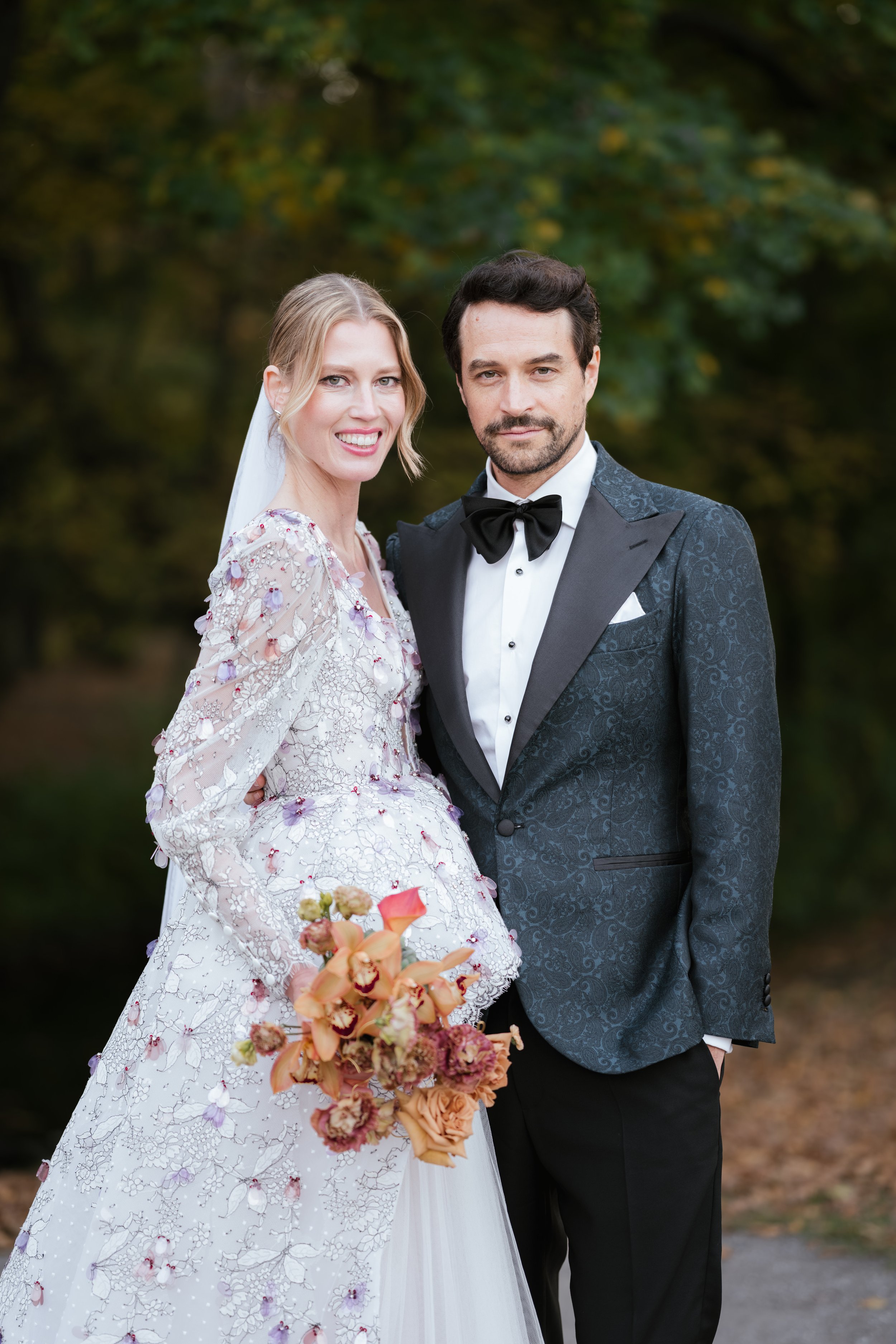 A bride and groom standing outdoors, dressed in wedding attire, smiling and posing for a photo with green trees in the background.