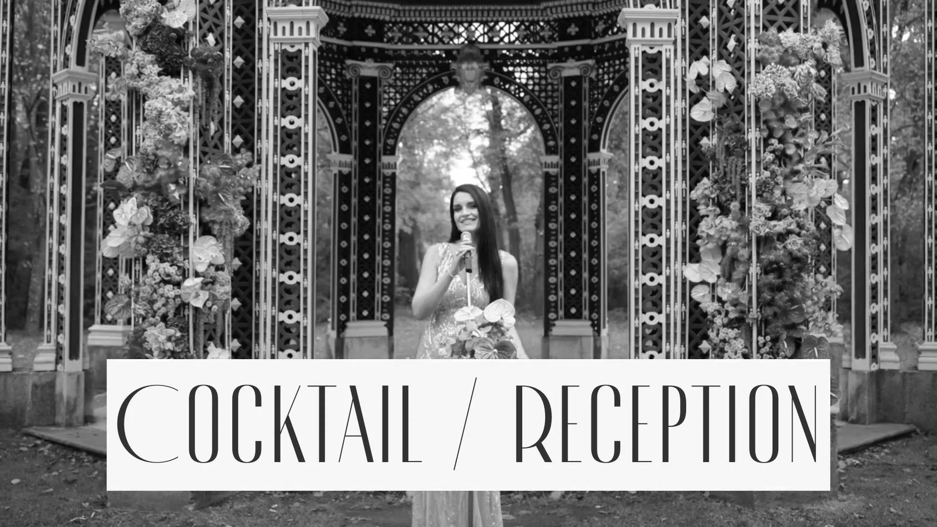 A woman standing in a floral decorated gazebo outdoors, holding a microphone, with a sign in front that reads "Cocktail / Reception".