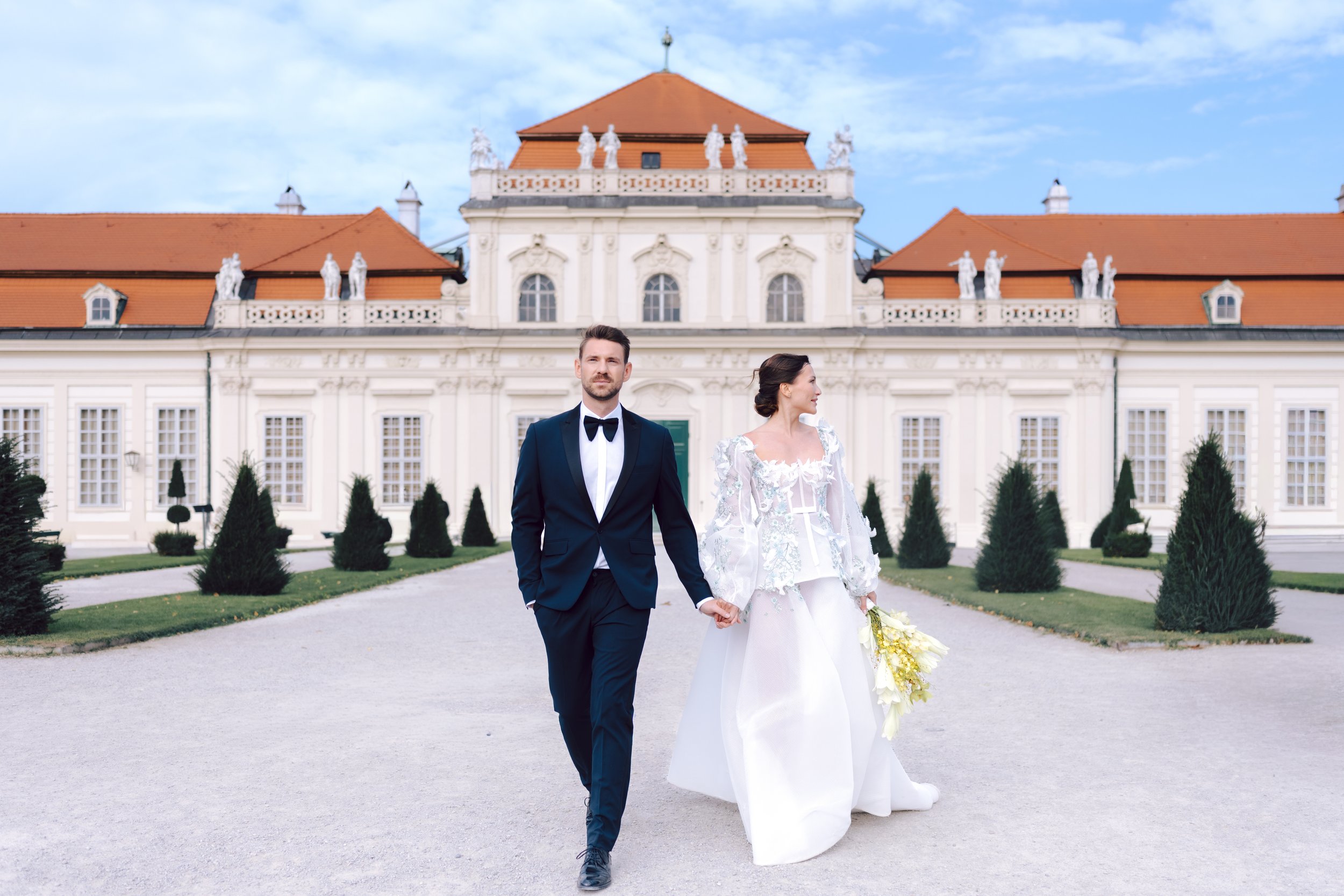 A bride and groom walking hand in hand in front of a grand, historic building with orange rooftops and decorative statues, on a clear day.