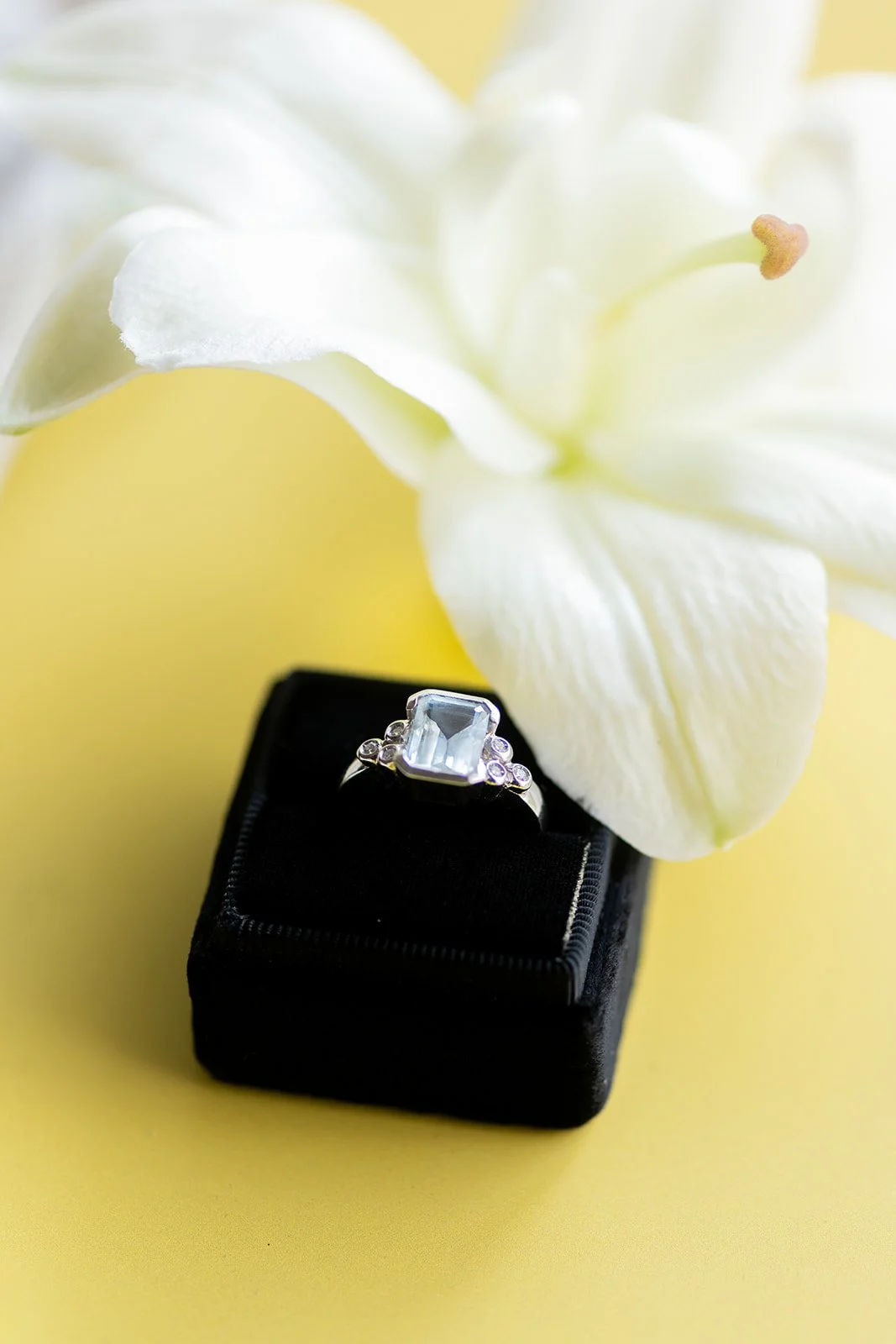 A silver ring with a large rectangular gemstone sits on a small black jewelry box. A white lily flower with soft petals is positioned behind the ring, against a yellow background.