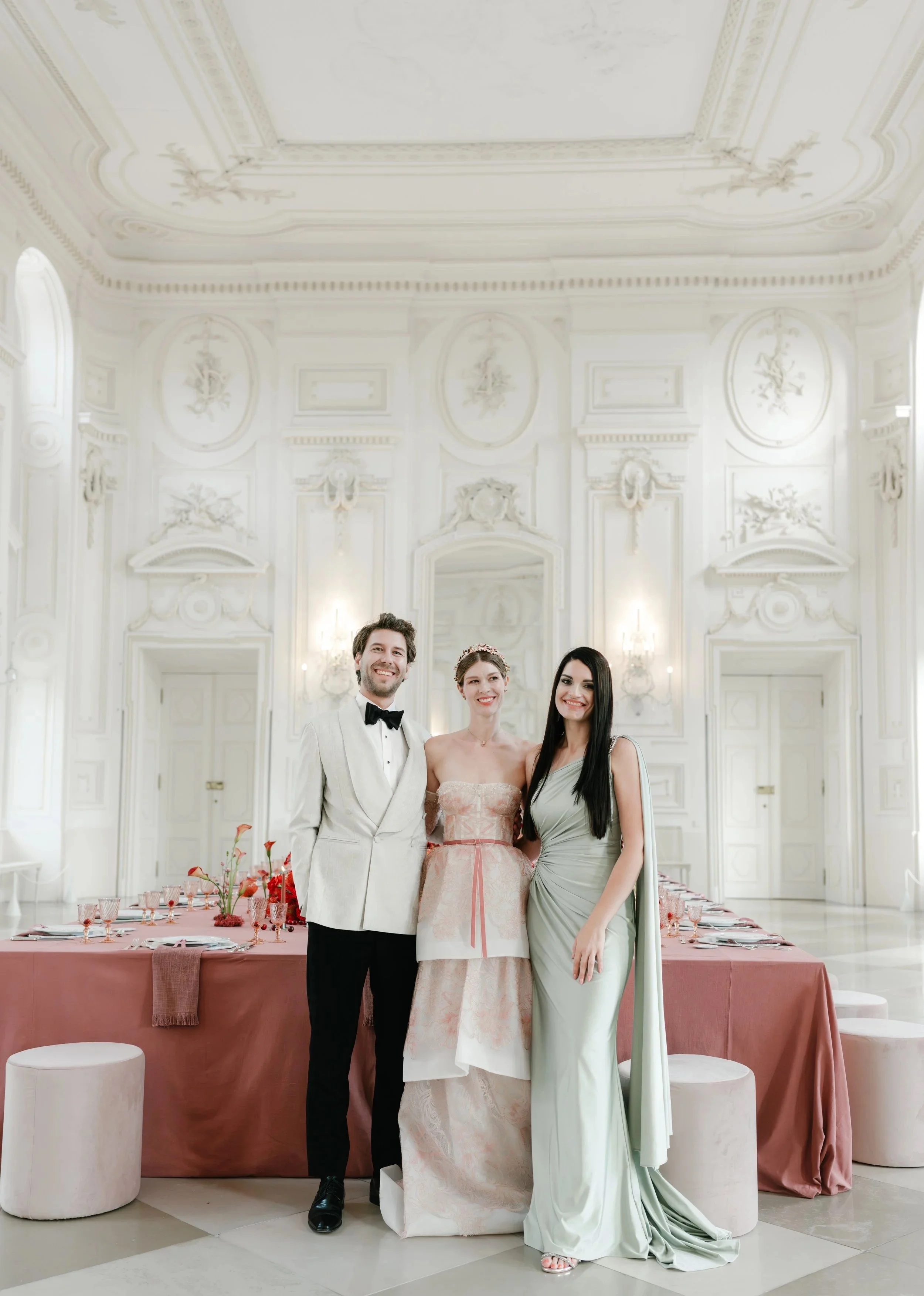 Three people in formal attire standing together in a luxurious, white ornate ballroom with elegant wall and ceiling decorations, and a long table set with pink tablecloth and floral arrangements in the background.