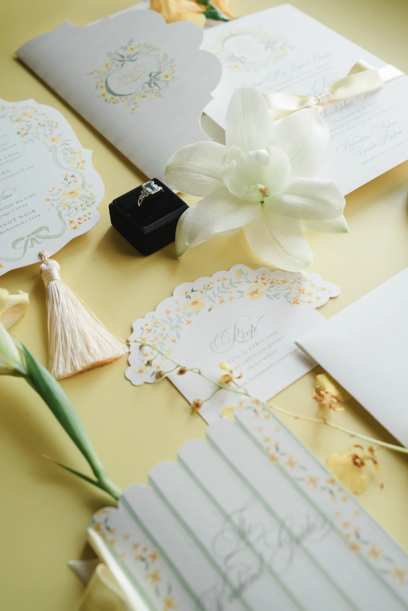 Wedding invitations, a white orchid flower, a ring in a black box, and decorative items on a yellow surface.