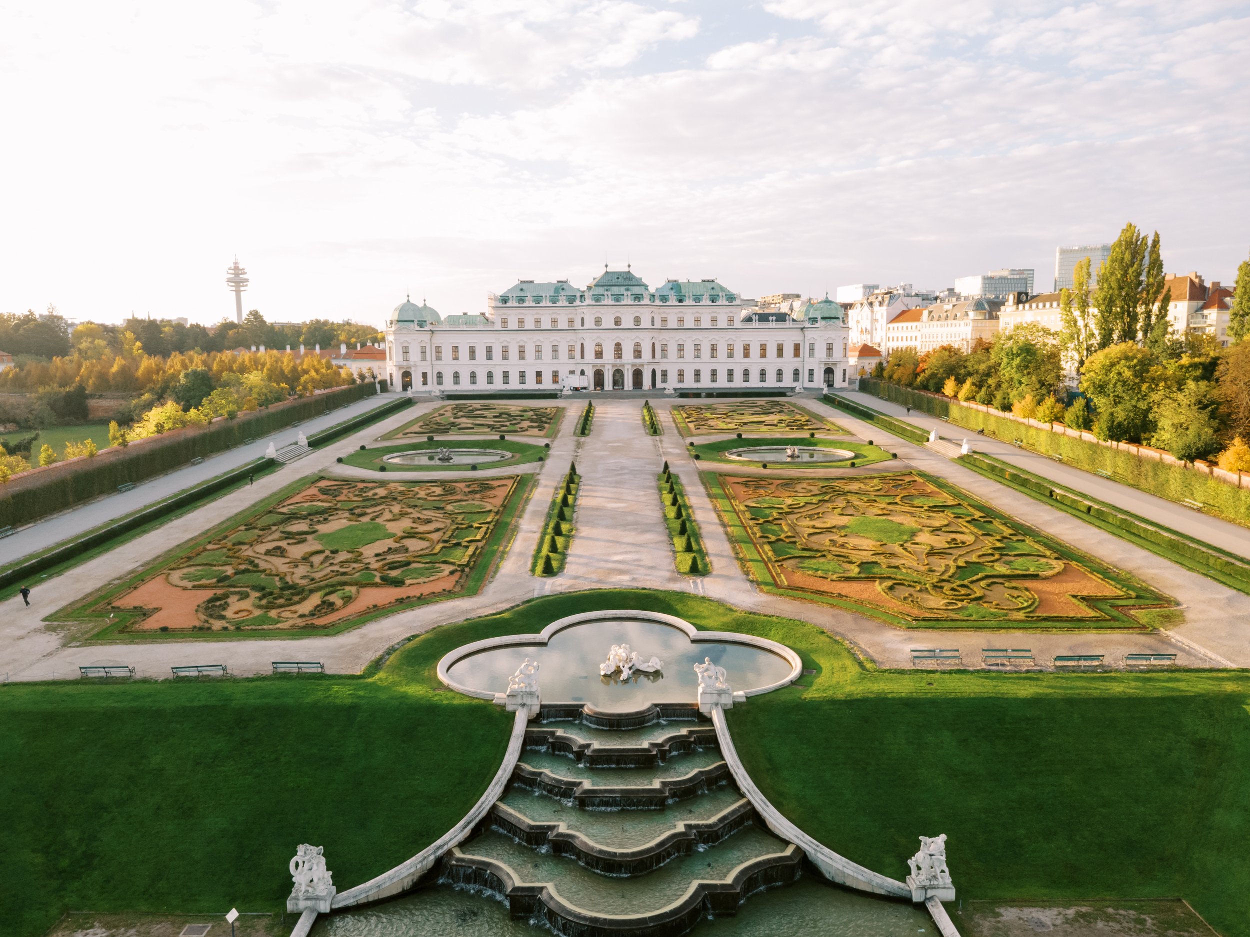 A grand historic building with elaborate garden landscaping in front, featuring symmetrical flowerbeds, fountains, and statues. This is Belvedere Castle in Vienna. Photo taken by Sotiris Tsakanikas.