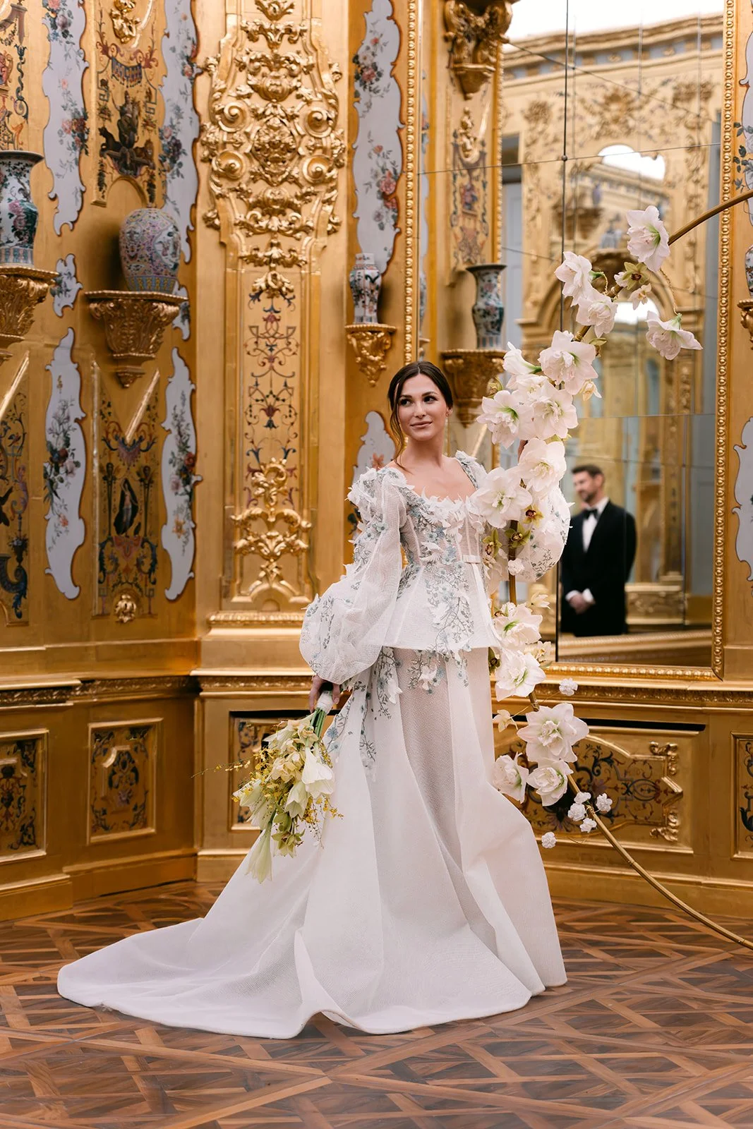 A woman in a wedding dress holding a bouquet of flowers before the civil ceremony or symbolic ceremony, standing in a luxurious, gold-adorned room with ornate moldings and a large mirror. A man in a tuxedo is visible in the background. Music by Anna.