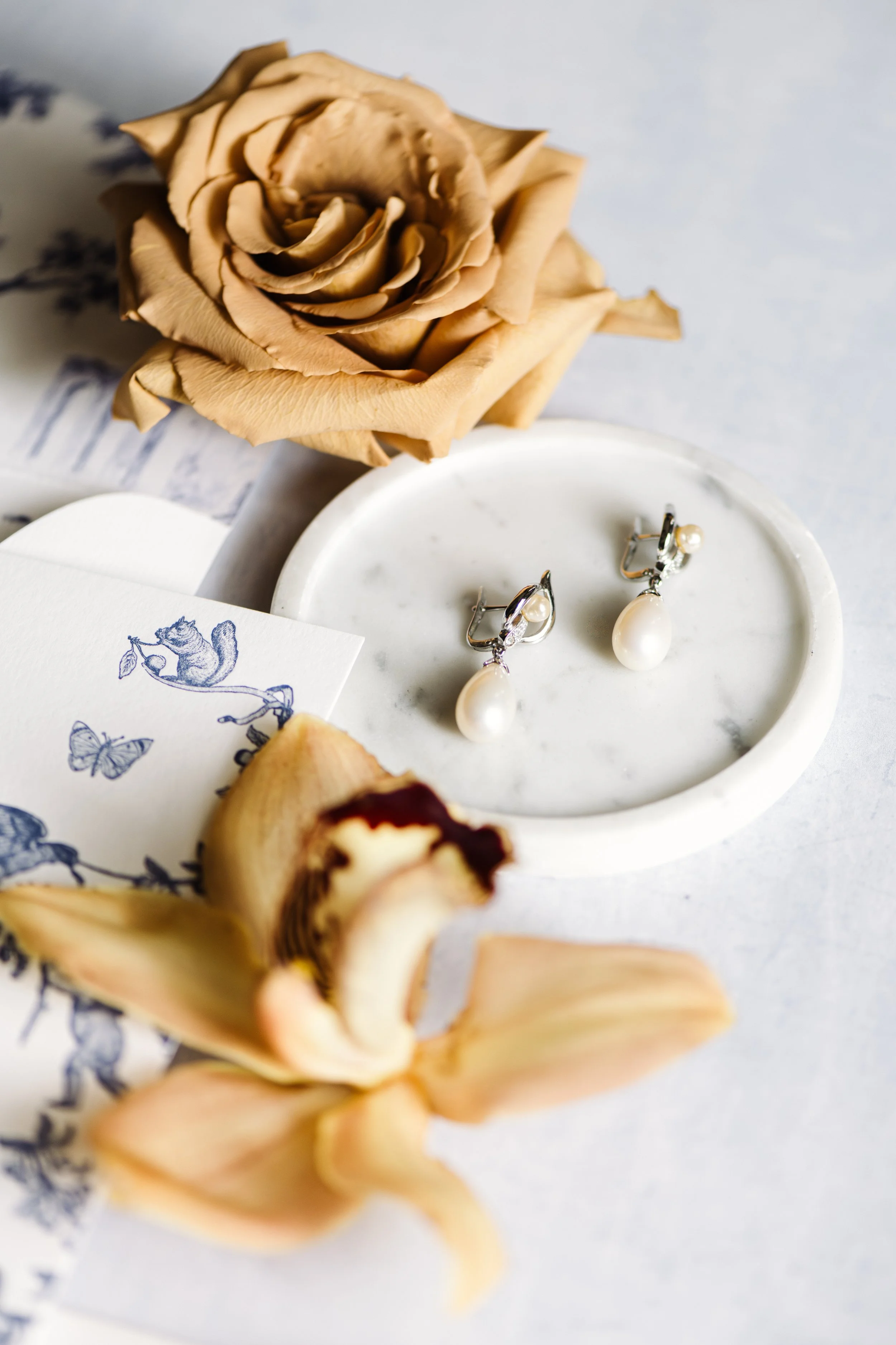 A pair of pearl drop earrings on a white marble dish, dried beige rose, and a partially opened banana with banana peel on a white surface.