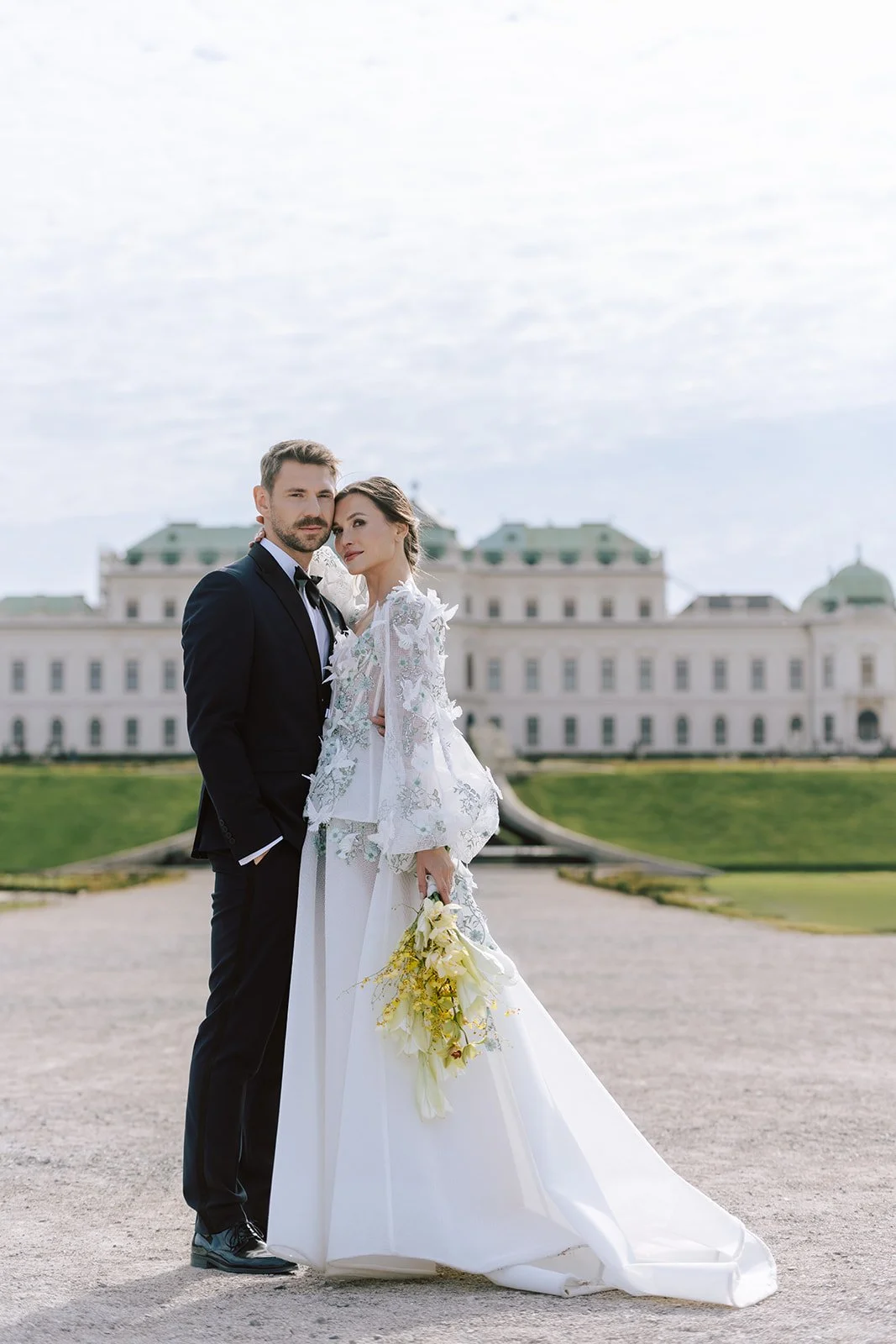 A bride and groom standing outside in front of a large white mansion, with the bride holding a bouquet of yellow and white flowers. The groom is dressed in a black tuxedo, and the bride is in a white wedding gown with lace details.