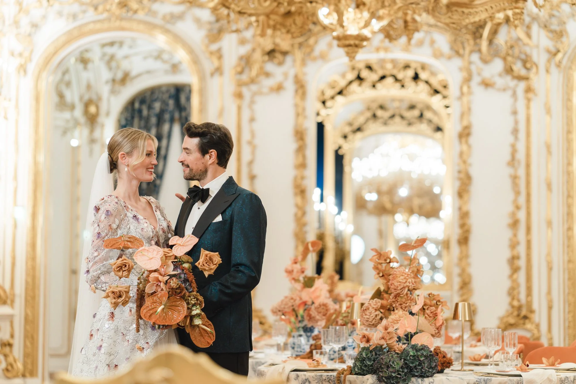 A bride and groom smiling at each other during their wedding reception in a luxurious, ornate hall at Palais Liechtenstein in Vienna, with gold accents and chandeliers, with a table decorated with pink and peach flowers. Photo by Sotirios Tsakanikas.