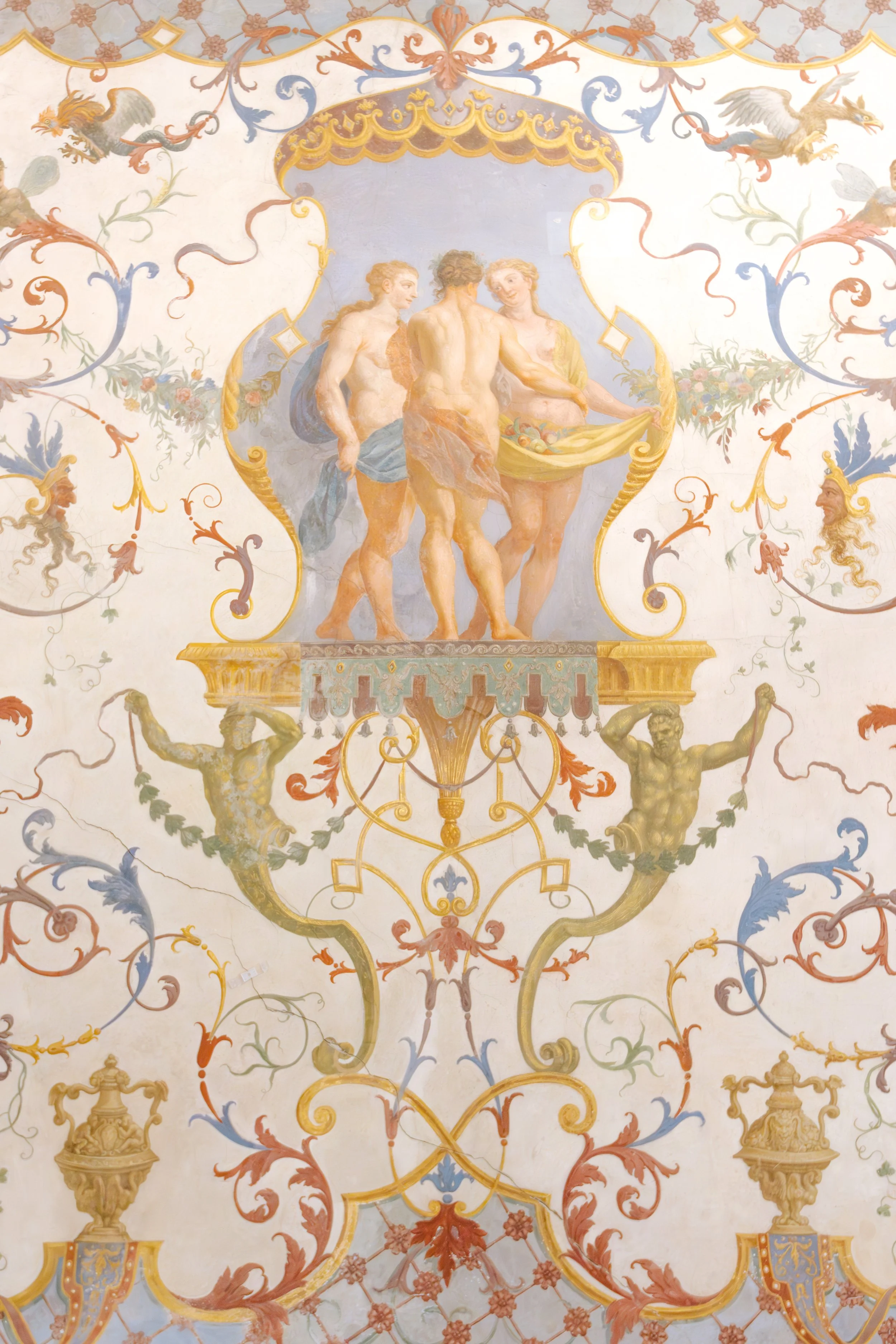 Fresco painting of three mythological figures, one holding a basket of fruits, with ornate decorative design including scrolls, floral motifs, and urns.