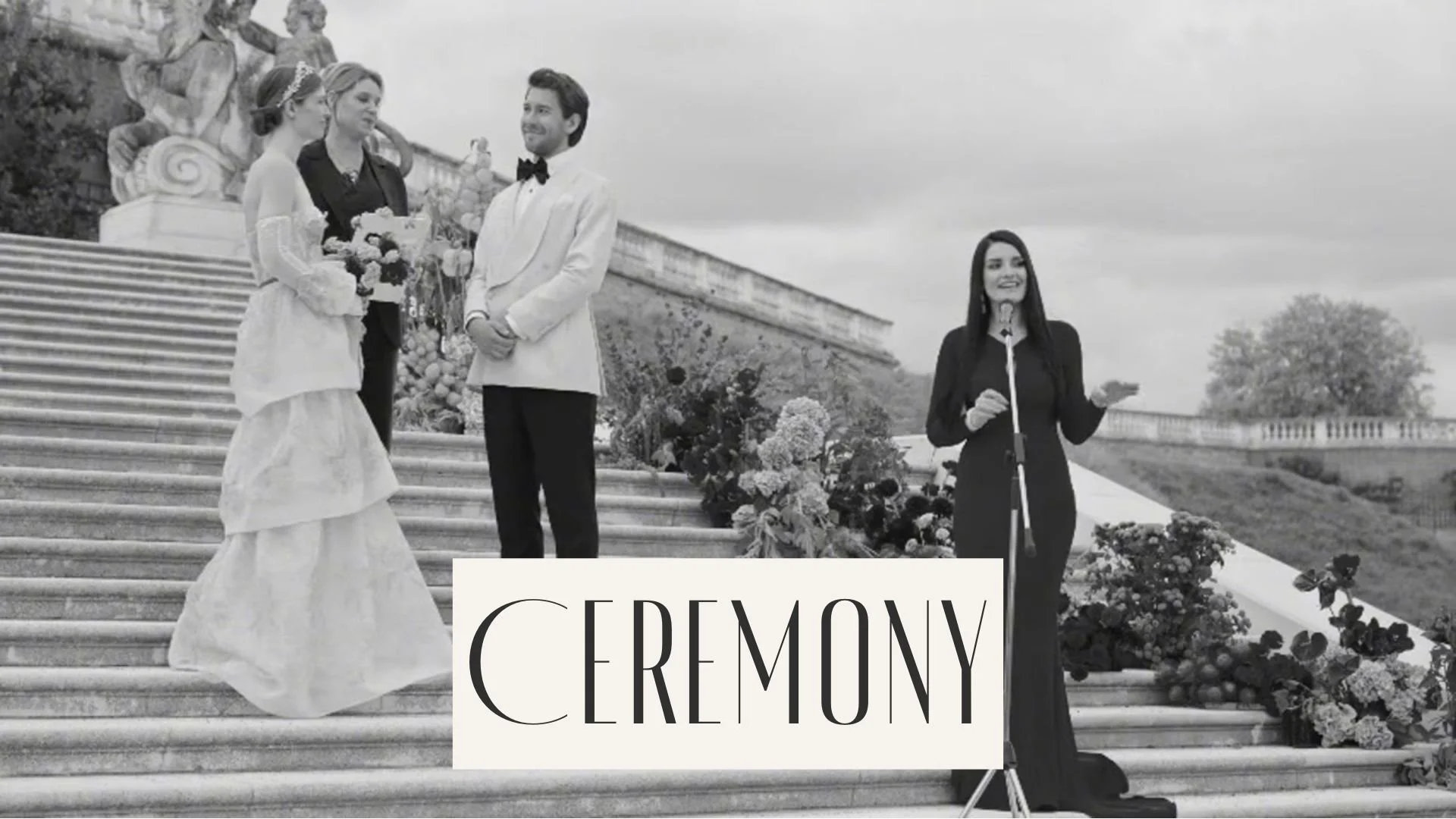 A black and white photo of a wedding ceremony on steps with a bride holding a bouquet, a groom in a tuxedo, and two women standing nearby, one speaking at a microphone.