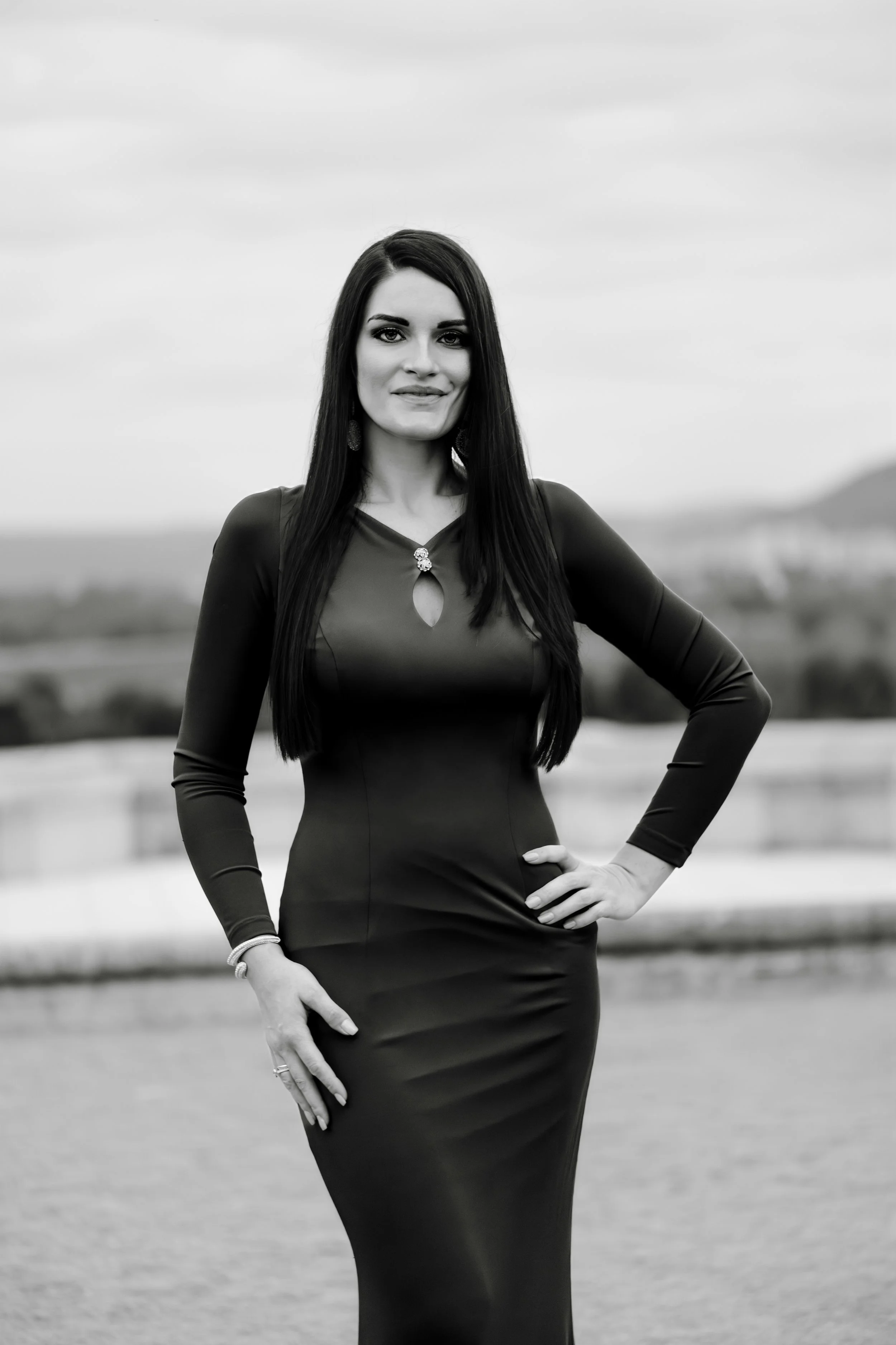 A woman with long dark hair, wearing a long-sleeved, form-fitting dress, standing outdoors with a blurred landscape background.