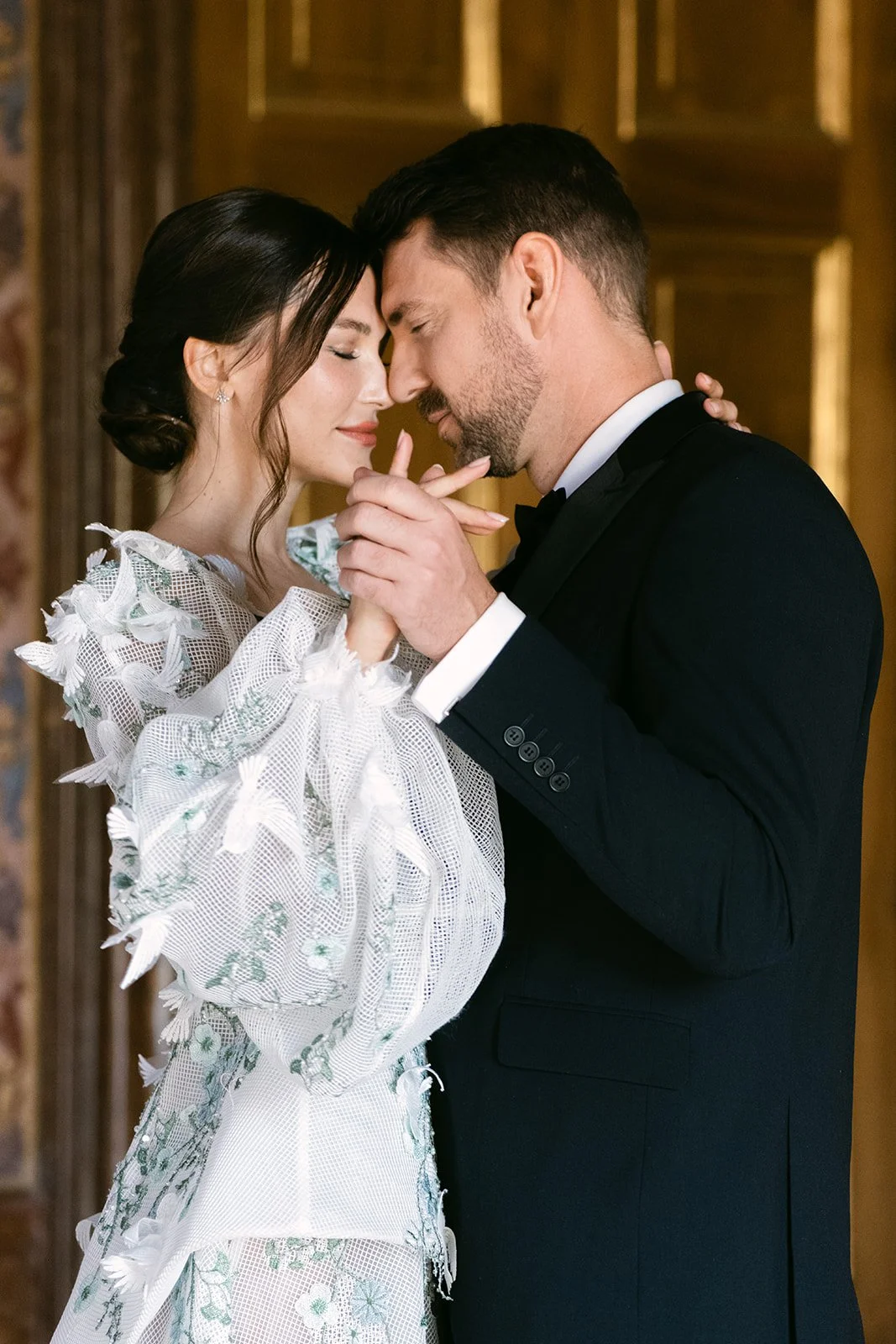A bride and groom share a tender moment with foreheads touching and eyes closed, holding hands in a warmly lit room.