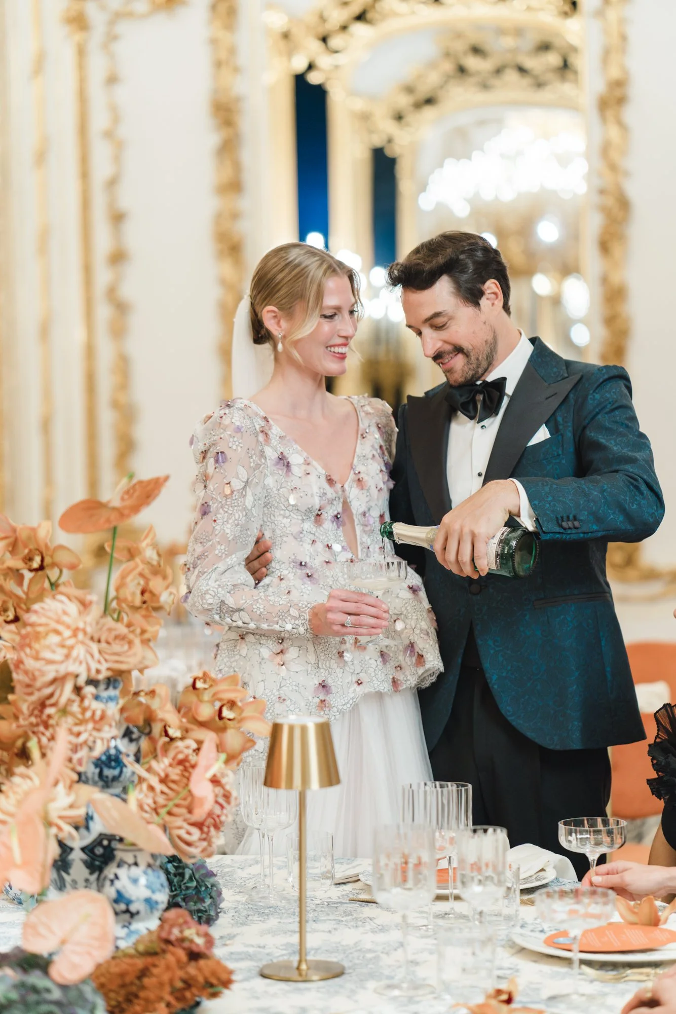 A bride and groom at a wedding reception, the groom is pouring champagne into a glass while the bride smiles. The setting is elegant with ornate gold decorations and floral arrangements.
