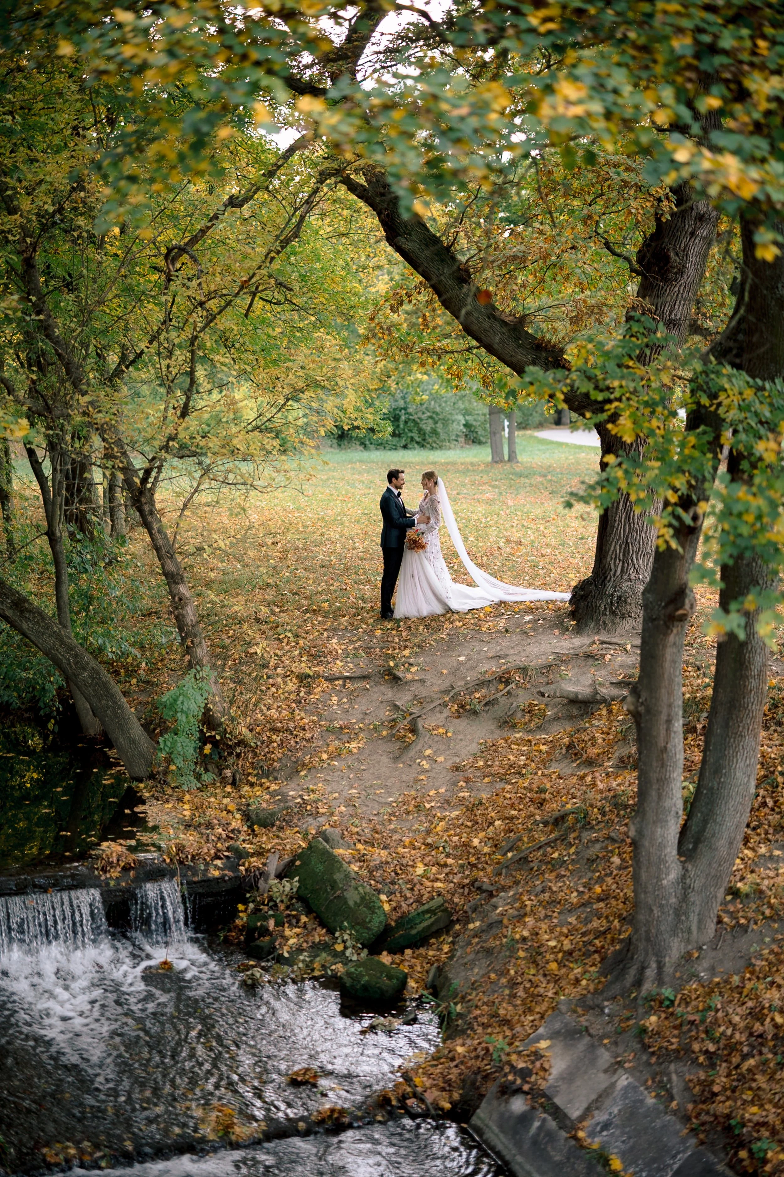 A bride and groom stand facing each other under large trees in an autumn park, holding hands, with a small stream and fallen leaves covering the ground.