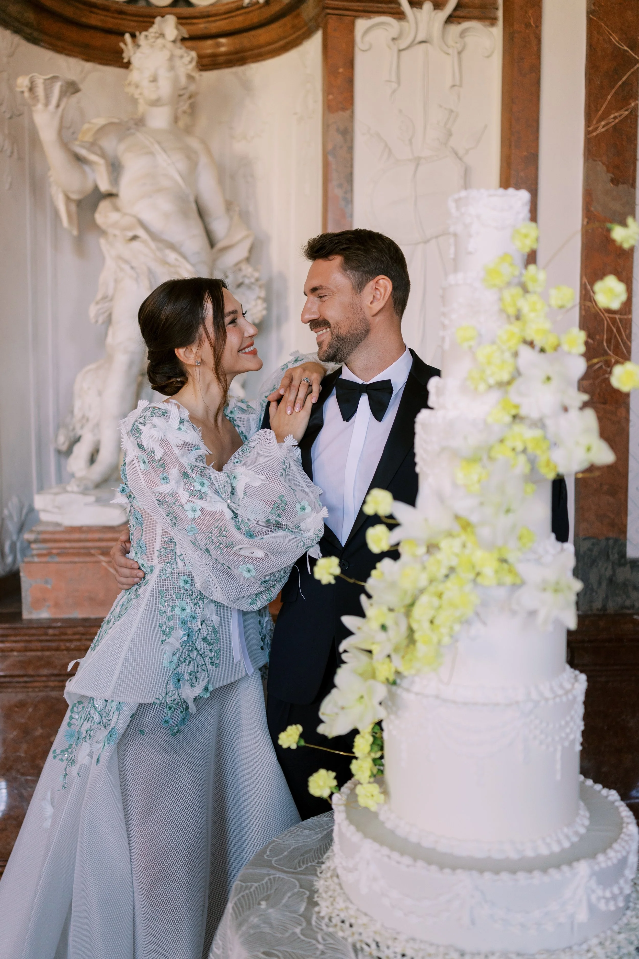 A bride and groom smiling at each other in a romantic way at their wedding reception with a tall white floral white and yellow wedding cake in the foreground and a classical statue in the background. They got married in Belvedere Castle in Vienna.
