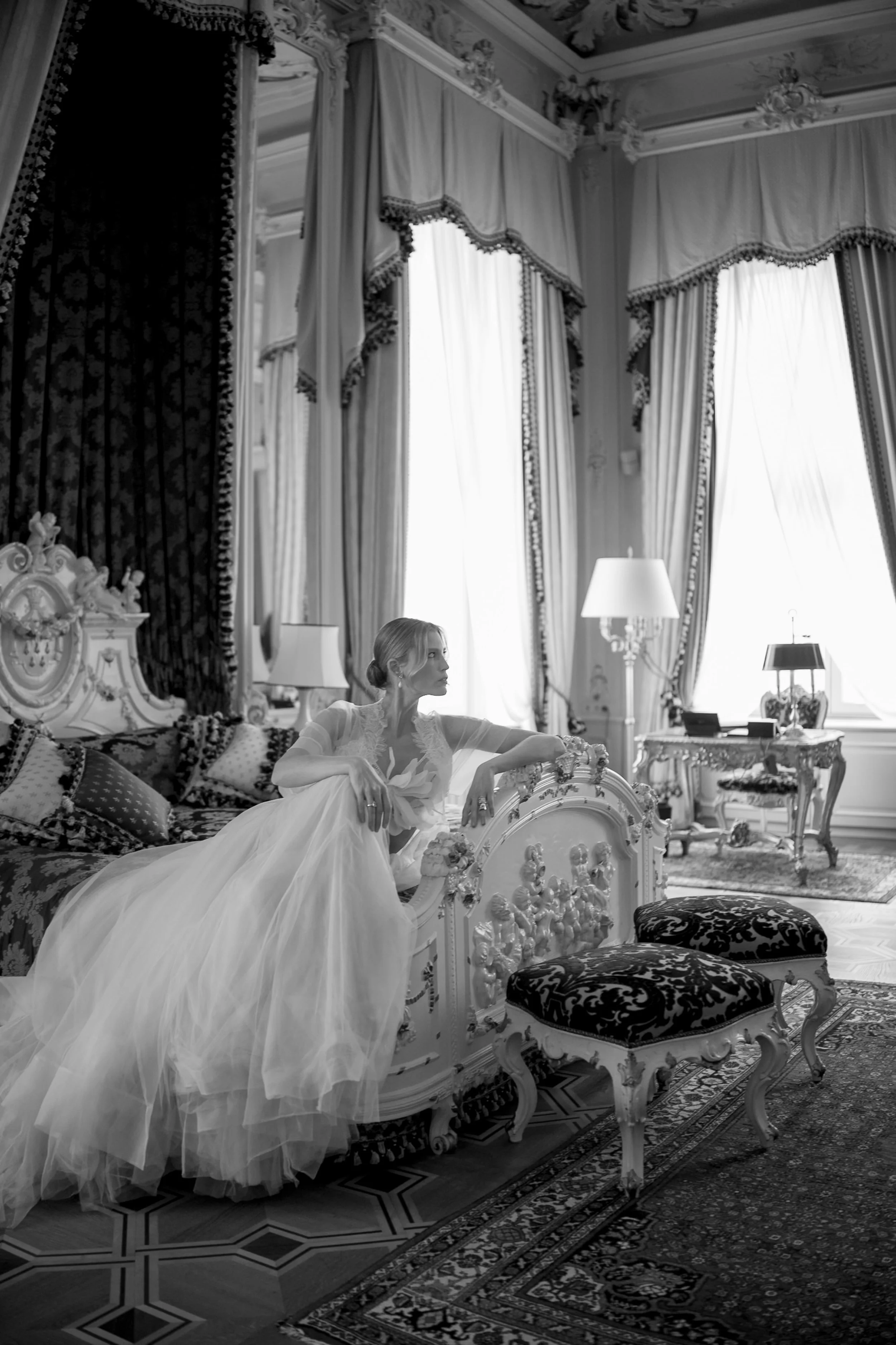 A woman in a beautiful luxurious wedding dress sitting on a vintage ornate bed in a luxurious, richly decorated hotel room in Vienna, with tall drapes, elaborate furniture, and soft lighting. Photo taken by Sotiris Tsakanikas.