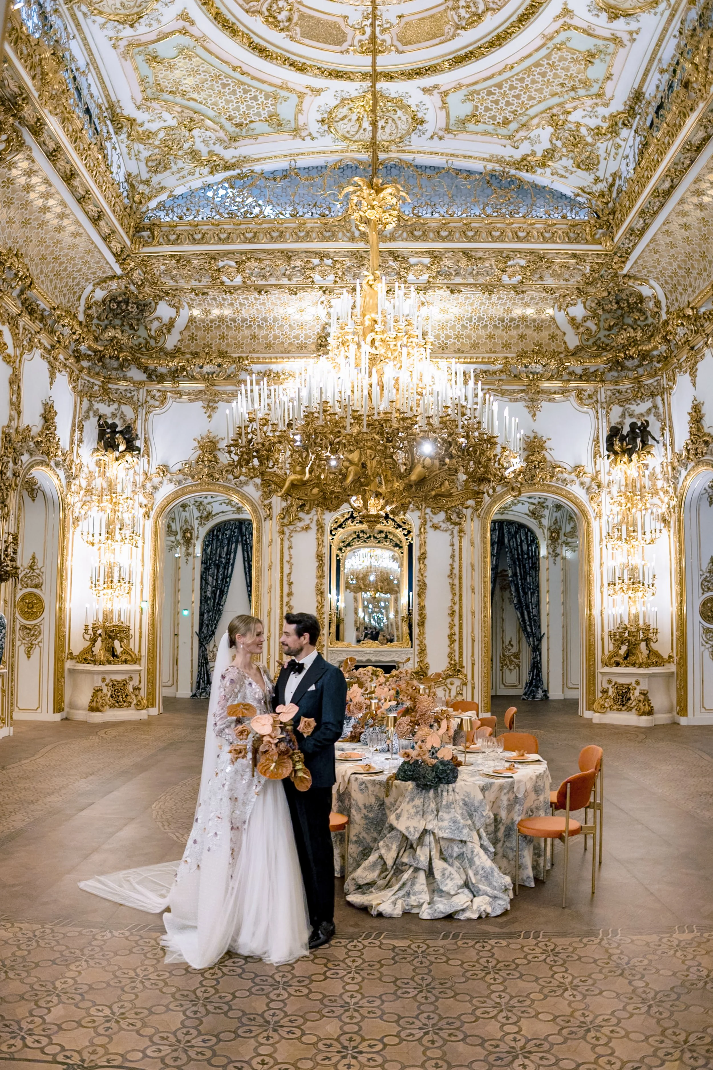 A bride and groom standing in an ornate, gold-accented ballroom with elaborate chandeliers and decorative wall moldings, near a decorated wedding reception table.