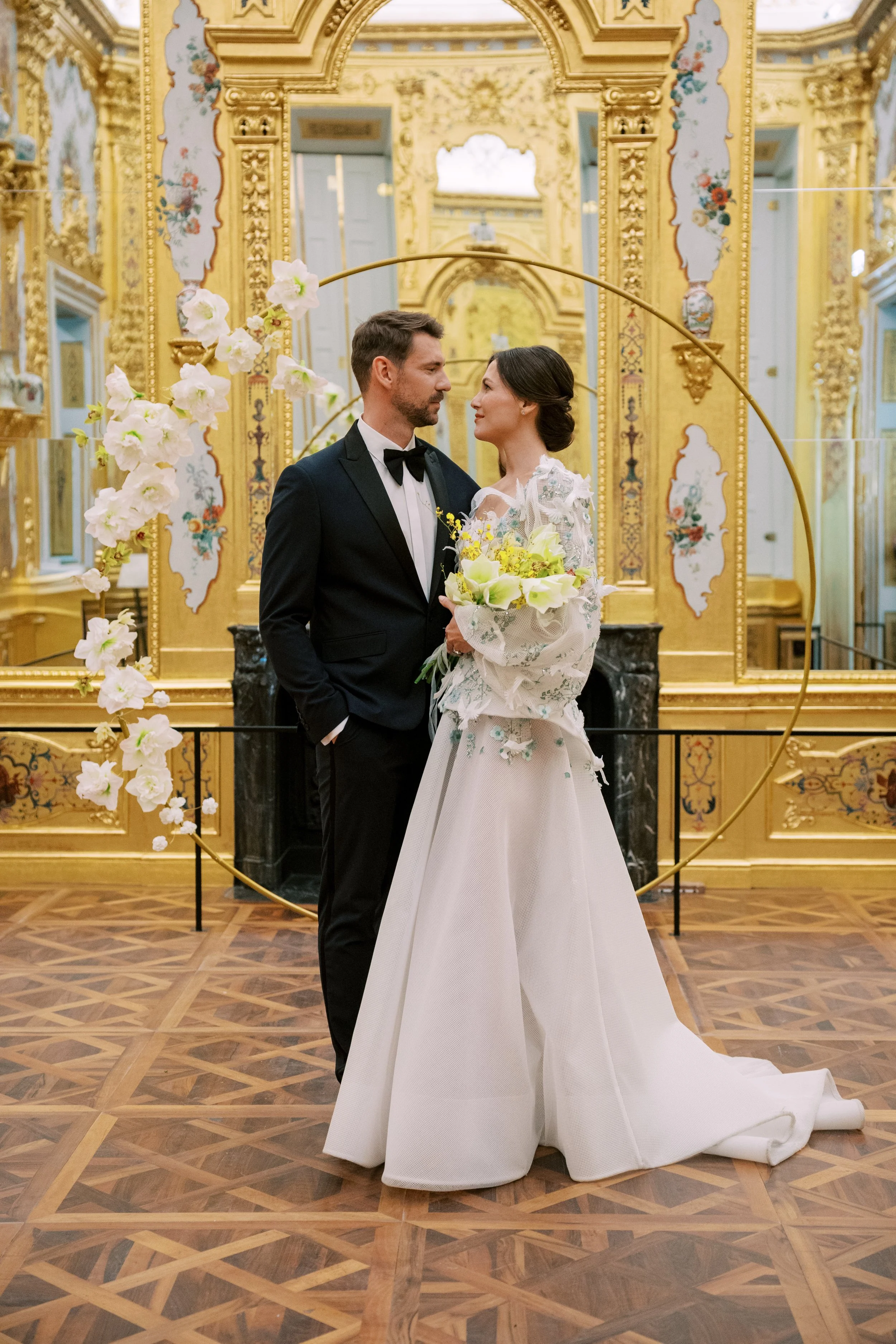 A bride and groom at their wedding ceremony inside a luxurious, ornate room with opulent golded walls, detailed wall decorations, surrounded by a circular white floral arrangement. This photo was taken by Sotiris Tsakanikas in Belvedere Castle,Vienna