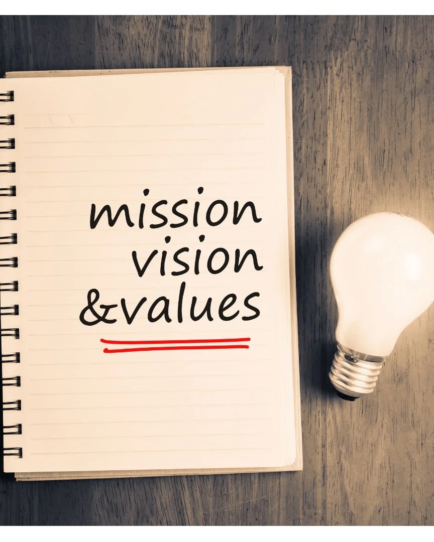Open notebook with lined pages displaying the words 'mission vision & values' with a red underline, placed on a wooden surface next to a light bulb.