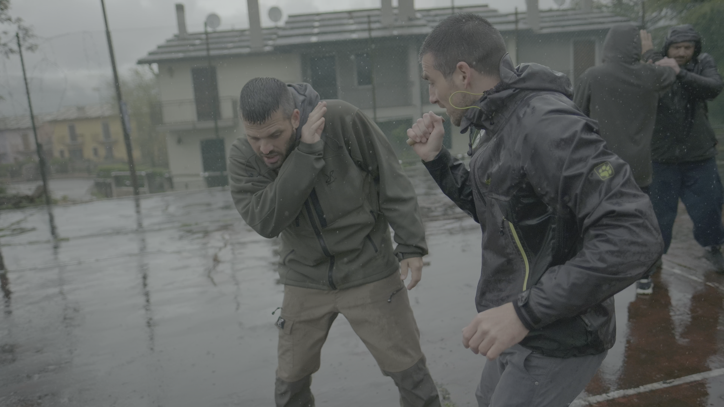 Zwei Männer kämpfen im Regen auf einem nassen Platz, während im Hintergrund weitere Personen Schutz suchen.