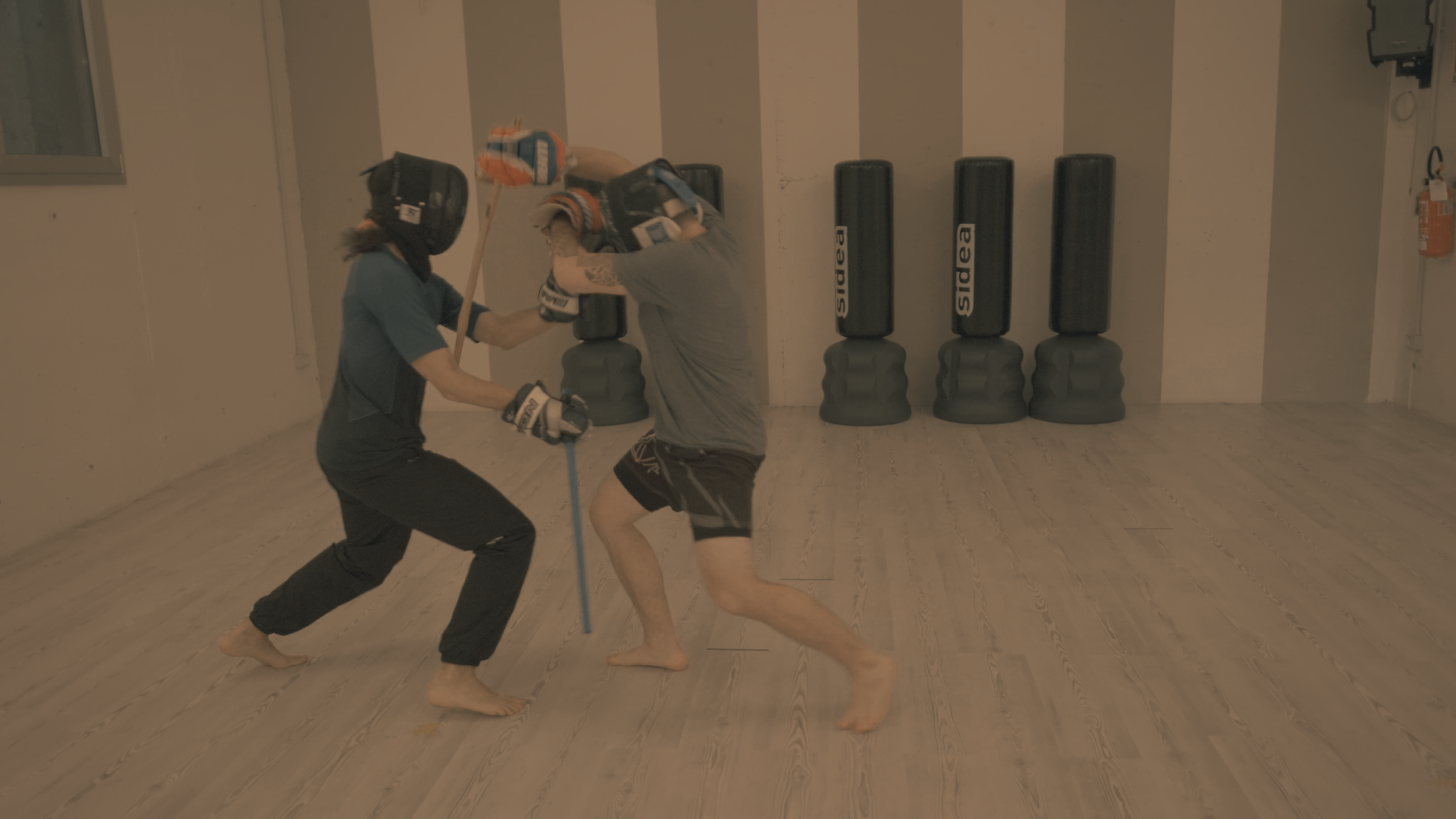 Zwei Menschen beim Kampfsport im Trainingsraum, einer trägt Schutzausrüstung und die andere hält einen Holzdorn