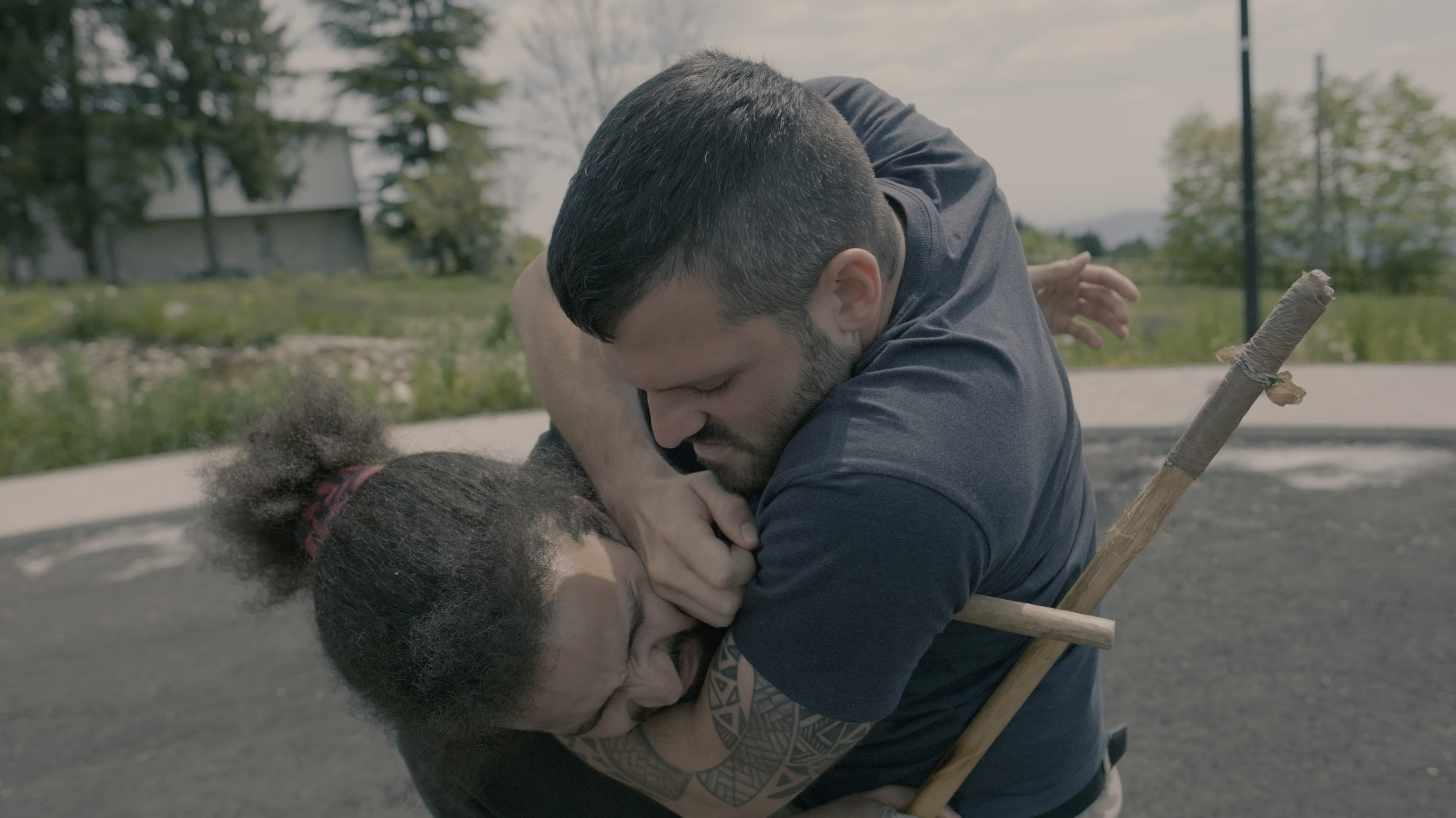 Zwei Männer in einem handgemachten Kampf, einer hält den anderen am Hals, im Hintergrund Bäume und ein Gebäude.