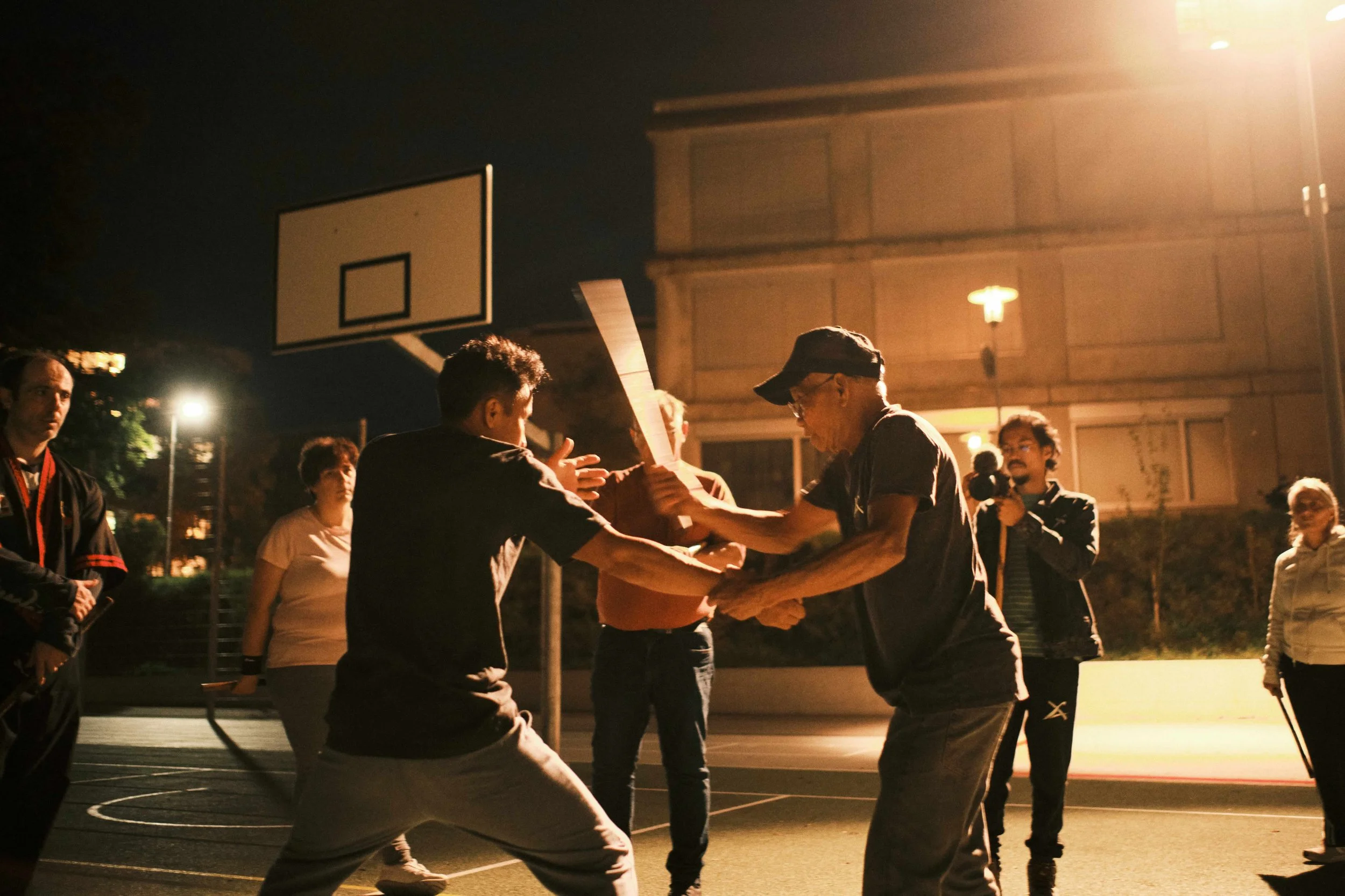 Menschen auf einem Basketballplatz bei einer nächtlichen Veranstaltung, einige halten sich an den Händen, andere beobachten.