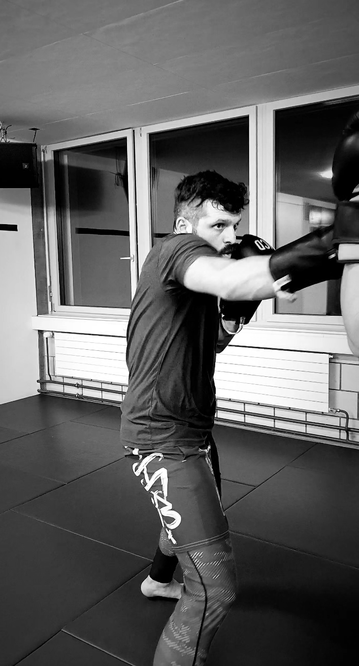 Zwei Männer bei einem Kampftraining, einer greift den anderen am Arm, in einem Raum mit Ziegelwänden.