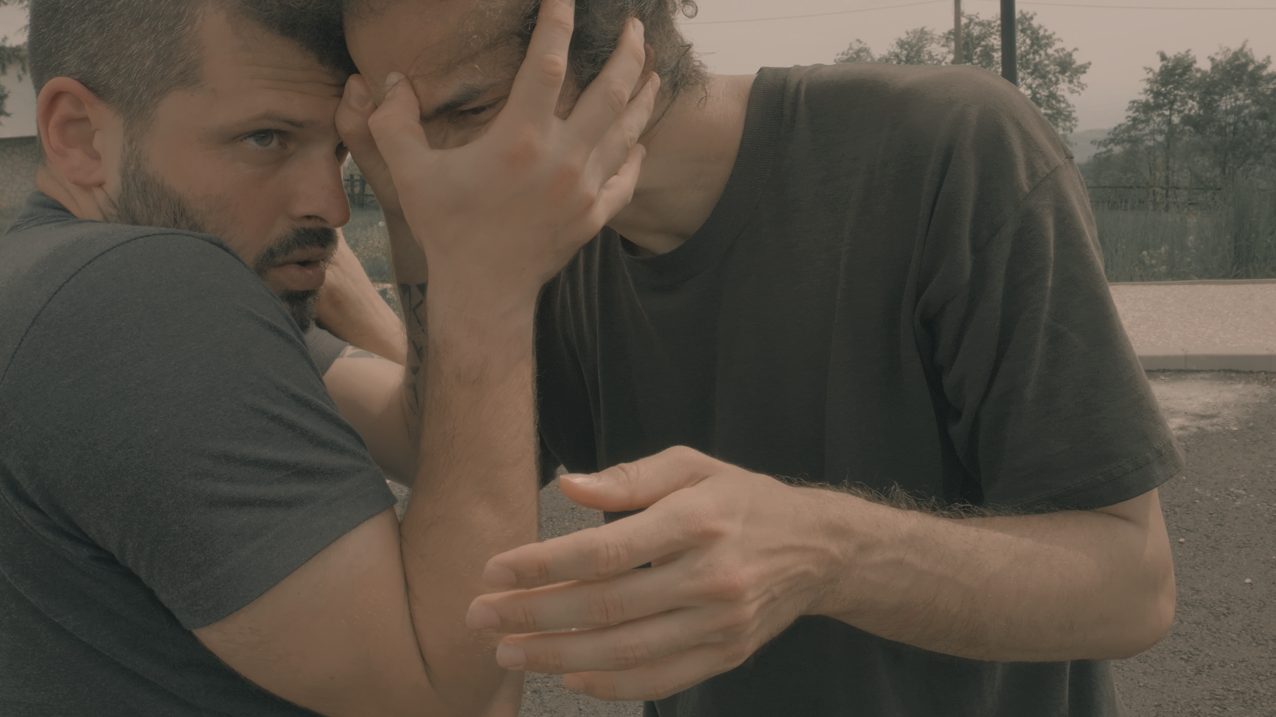 Zwei Männer sitzen eng umeinander, einer hält den Kopf des anderen in den Händen, beide scheinen emotional betroffen, draußen mit Bäumen im Hintergrund.