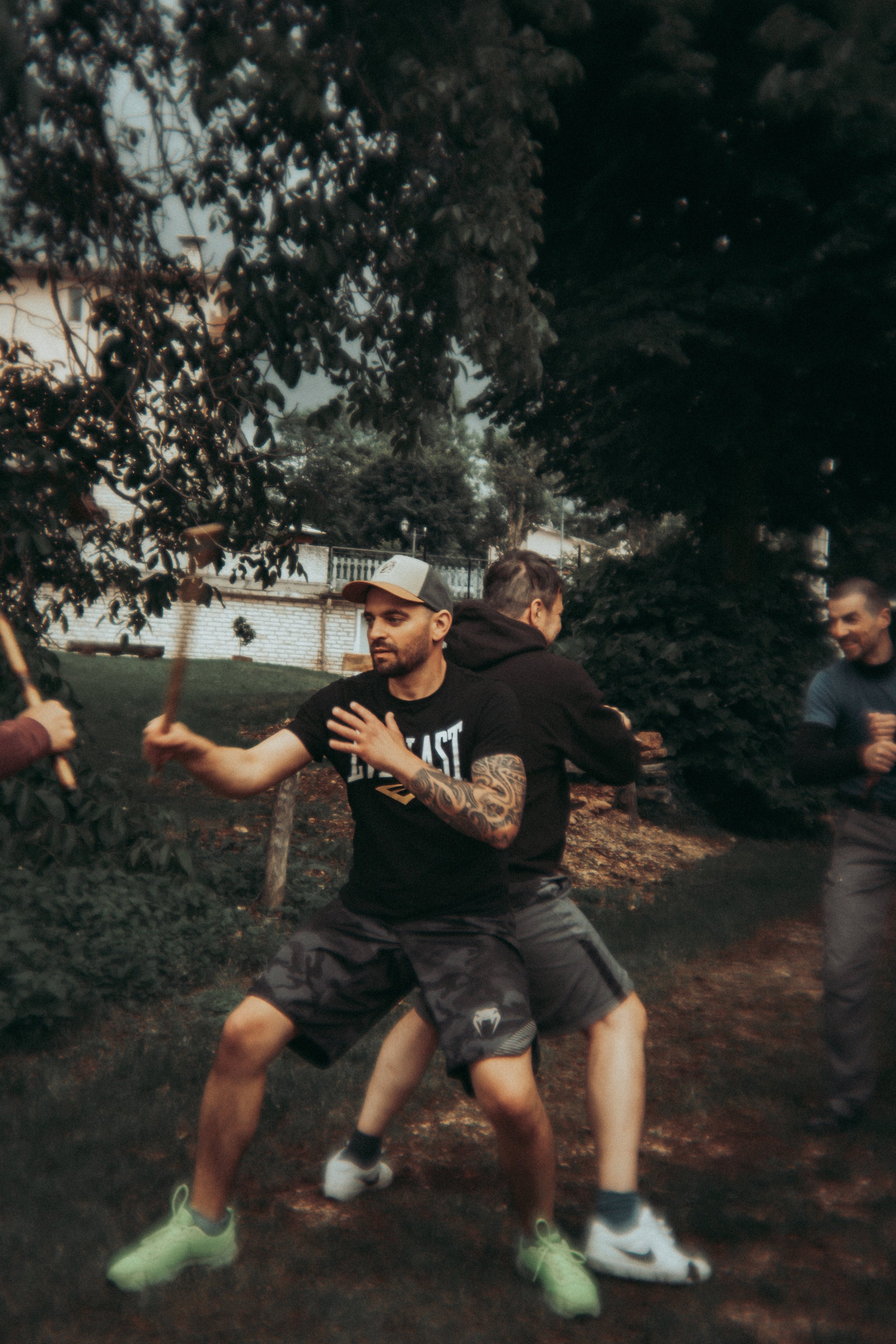 Zwei Männer trainieren im Freien mit Kampfsporttechnik, eines hat Tattoos auf dem Arm, trägt eine schwarze T-Shirt und Shorts, während der andere in dunklem T-Shirt und Shorts ist.