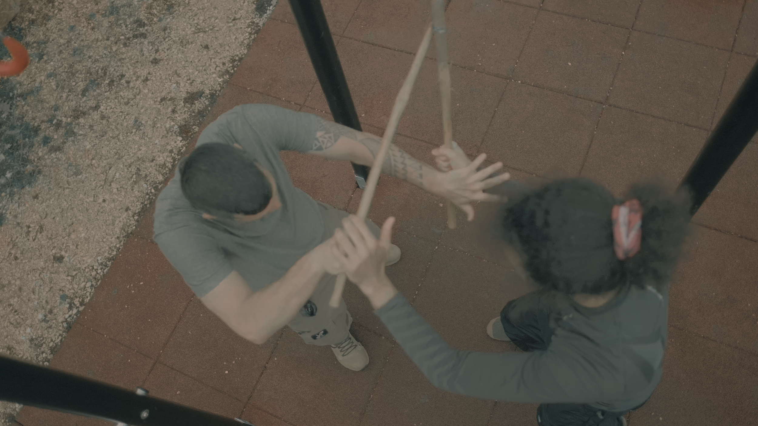 Zwei Menschen bei einem Handstand an einem Spielplatz. Sie stoßen sich gegenseitig die Hände ab, während sie um einen Schaukelsatz stehen.