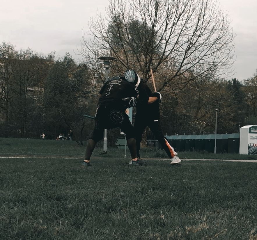 Zwei Personen führen eine Kampfsportübung im Freien aus, wobei sie in Kampfhaltung stehen, mit Stöcken in den Händen, in einem Park mit Bäumen im Hintergrund.
