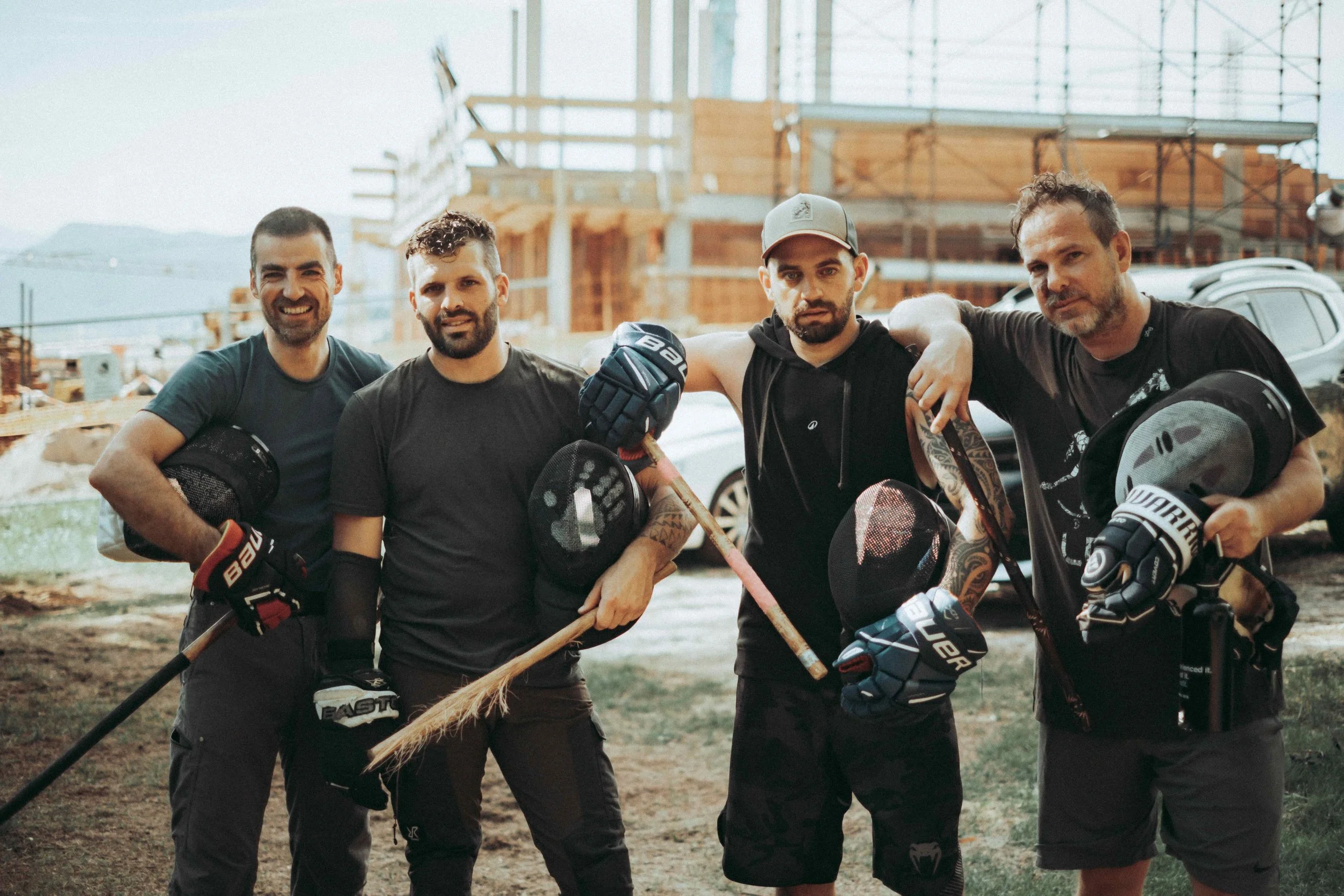 Fünf Männer in Eishockeyausrüstung posieren auf einer Baustelle, einige tragen Kellen, andere tragen Helme, im Hintergrund sind Baugerüste und ein Gebäude im Rohbau.