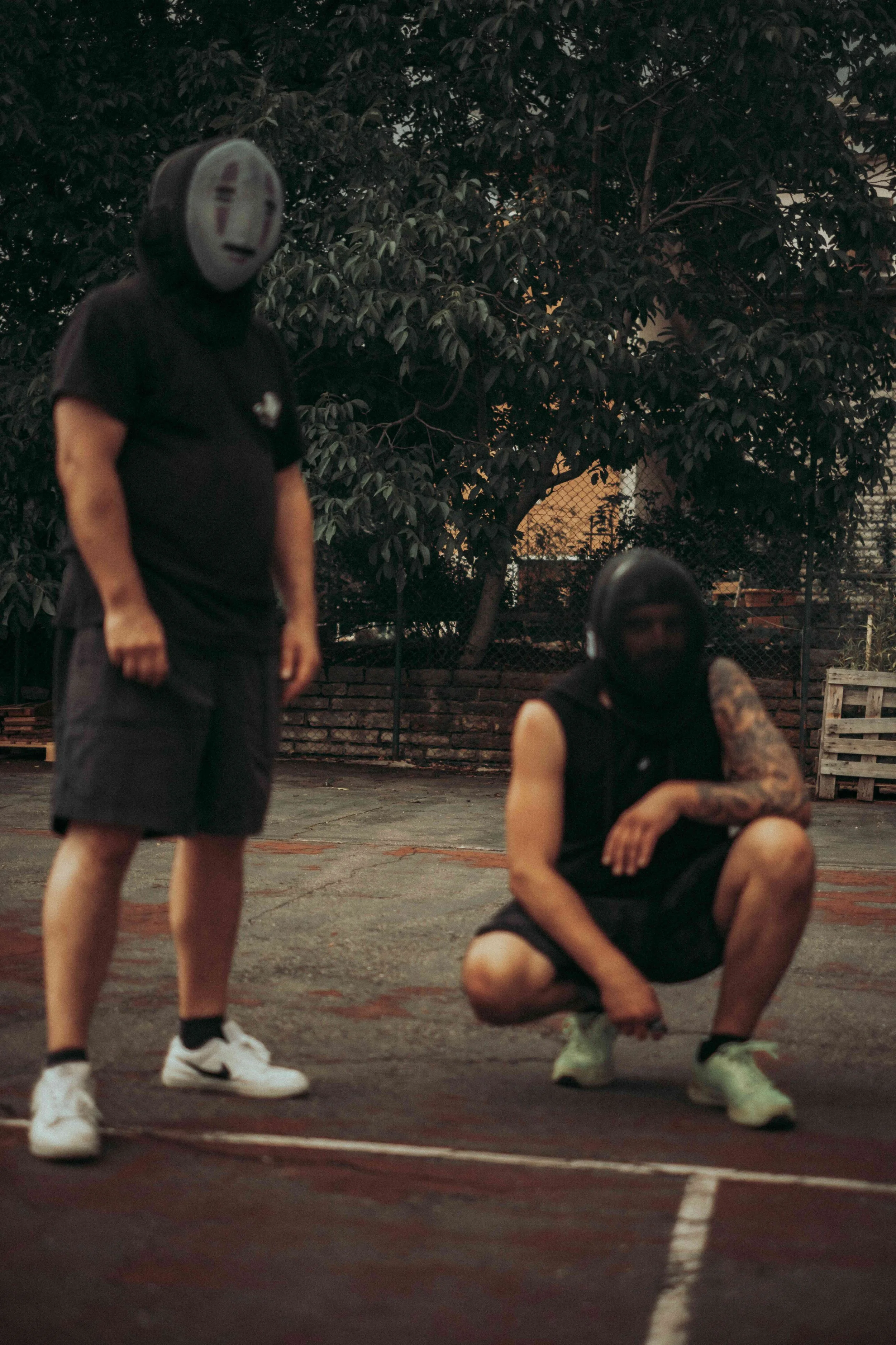 Zwei Männer mit schwarzen T-Shirts, einer trägt ein Totenkopf-Design und ein Masken mit einem lachenden Gesicht, posieren auf einem Tennisplatz. Einer steht, der andere sitzt auf dem Boden, umgeben von Bäumen