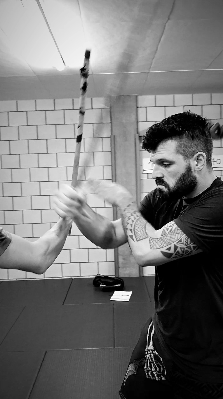 Zwei Männer bei einem Kampftraining, einer greift den anderen am Arm, in einem Raum mit Ziegelwänden.