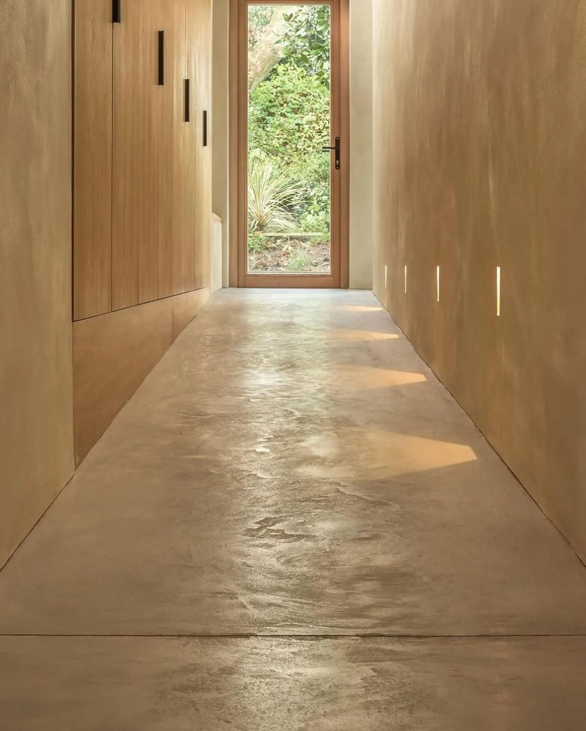 A minimalist hallway with wooden walls and a polished concrete floor, leading to a glass door with greenery outside.
