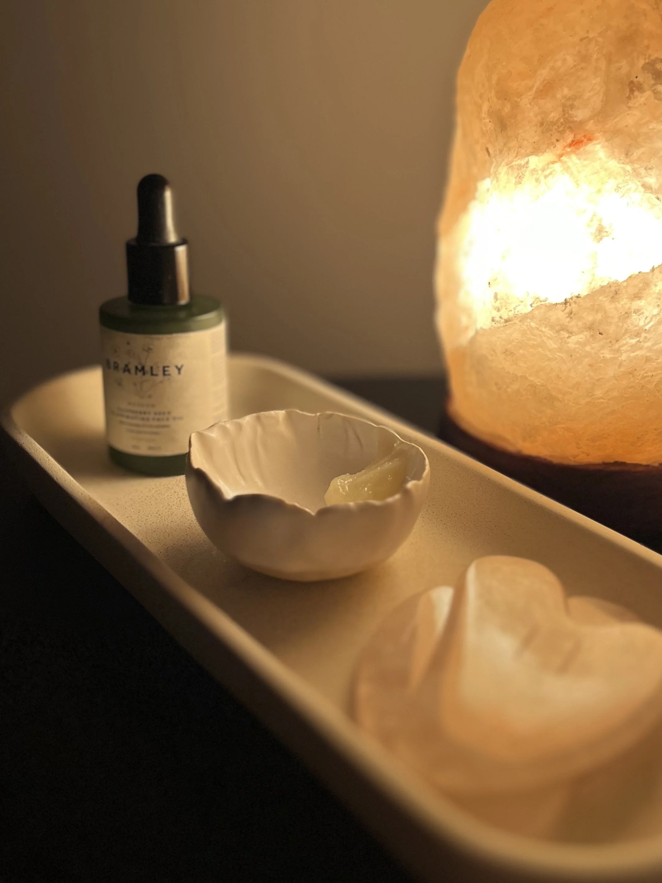 A salt lamp emitting a warm glow, a small bowl with a candle and some wax, a green dropper bottle, and a spoon with cream on a rectangular tray.